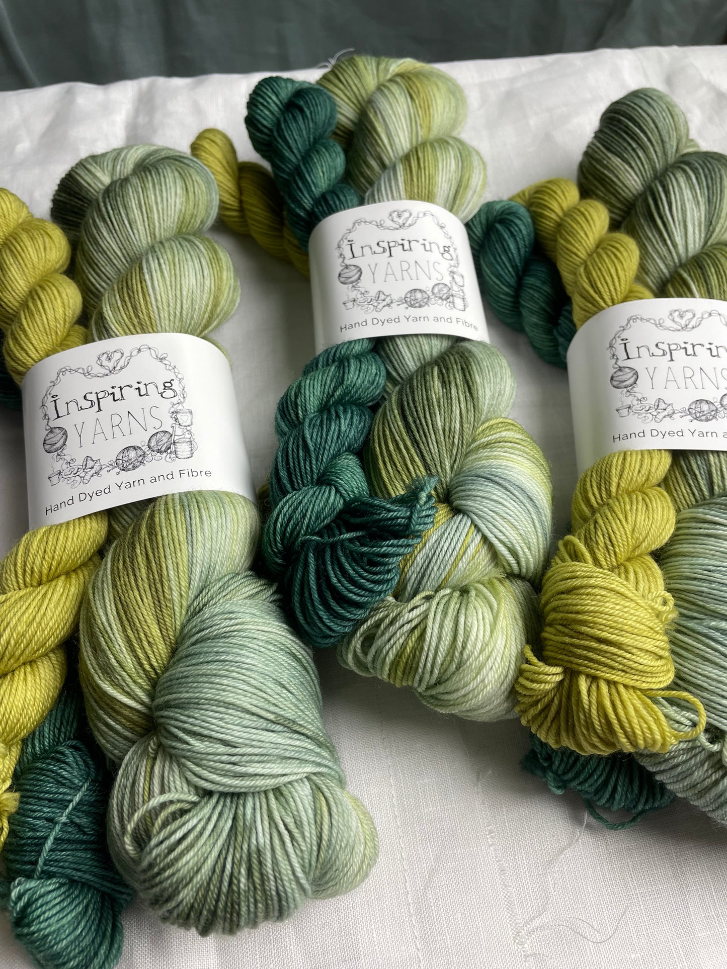 Three hand dyed yarn sock sets featuring large skein in green tones with a teal and a lime green mini skein labelled Inspiring Yarns. The background is white linen and some Christmas lights with pinecones, twigs and stars.