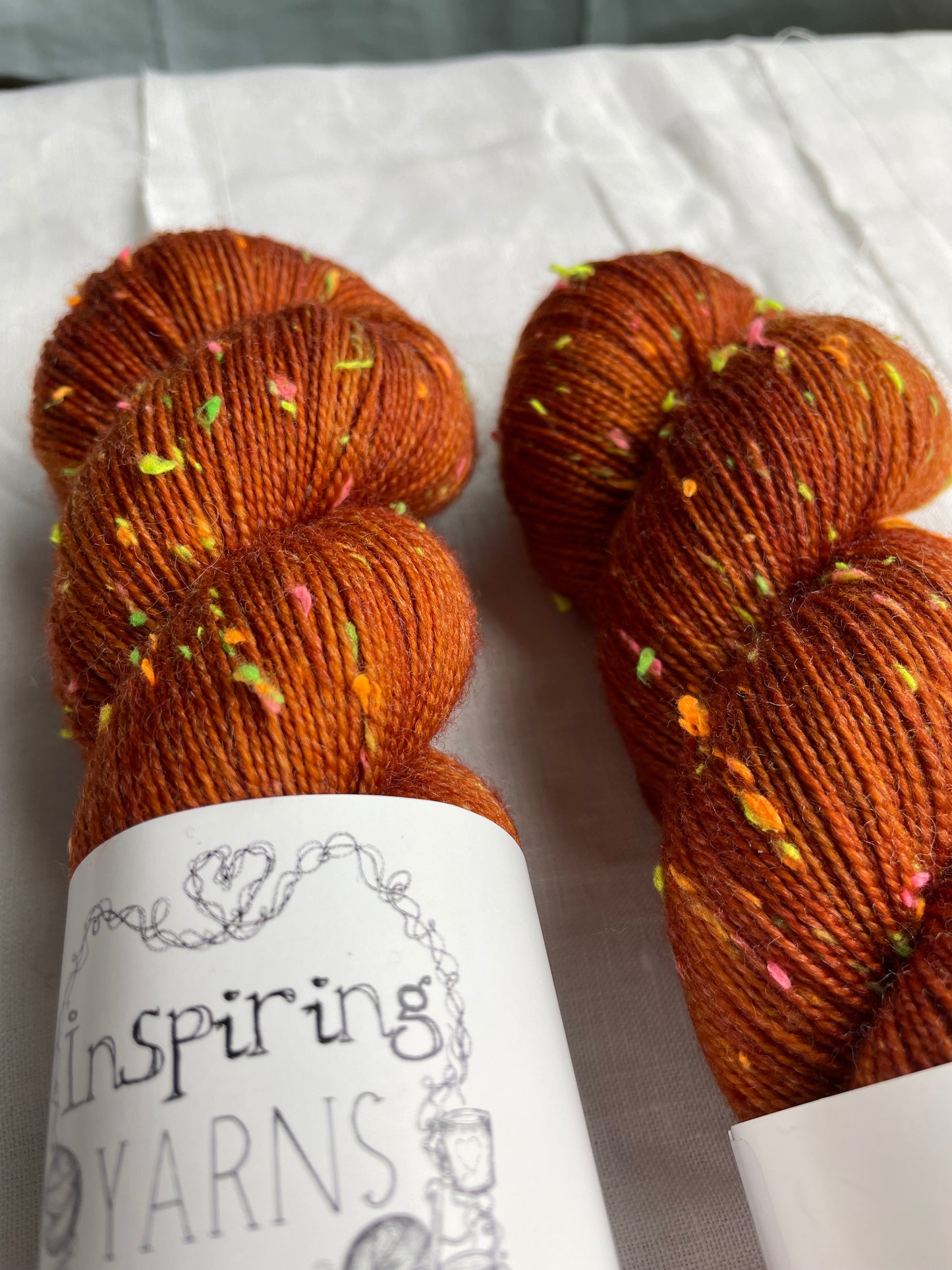 Close up of two skeins of tonal burnt orange coloured hand dyed Disco Donegal yarn branded Inspiring Yarns on a white linen background. The yarn has multi coloured neon neps.