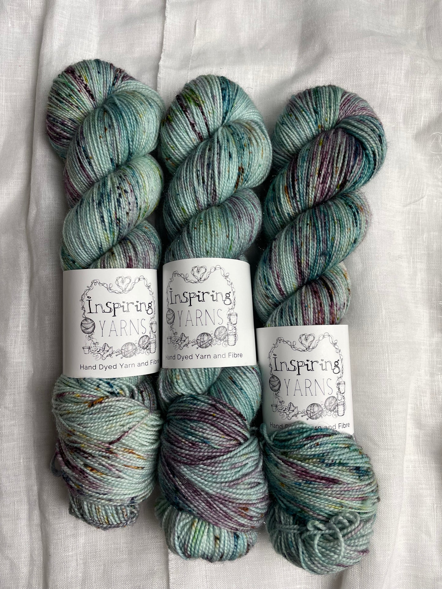 Three skeins of teal blue speckled Silver Sparkle Sock yarn labelled Inspiring Yarns on a white linen background.