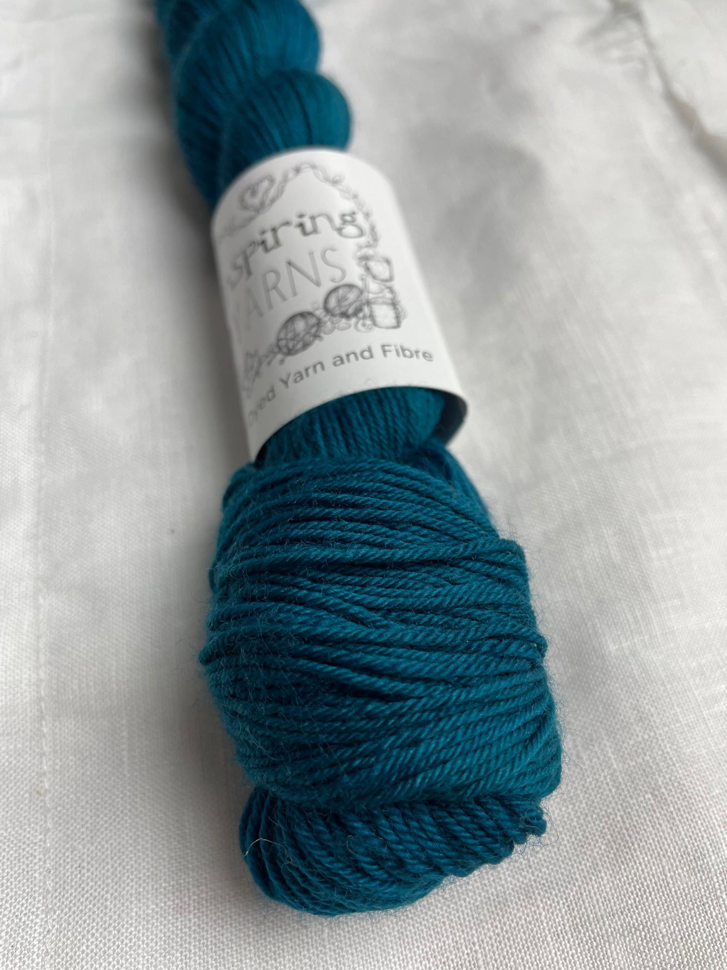 Close up of an intense teal blue coloured skeins of Merino Smooth Sock yarn branded Inspiring Yarns on a white linen fabric background.