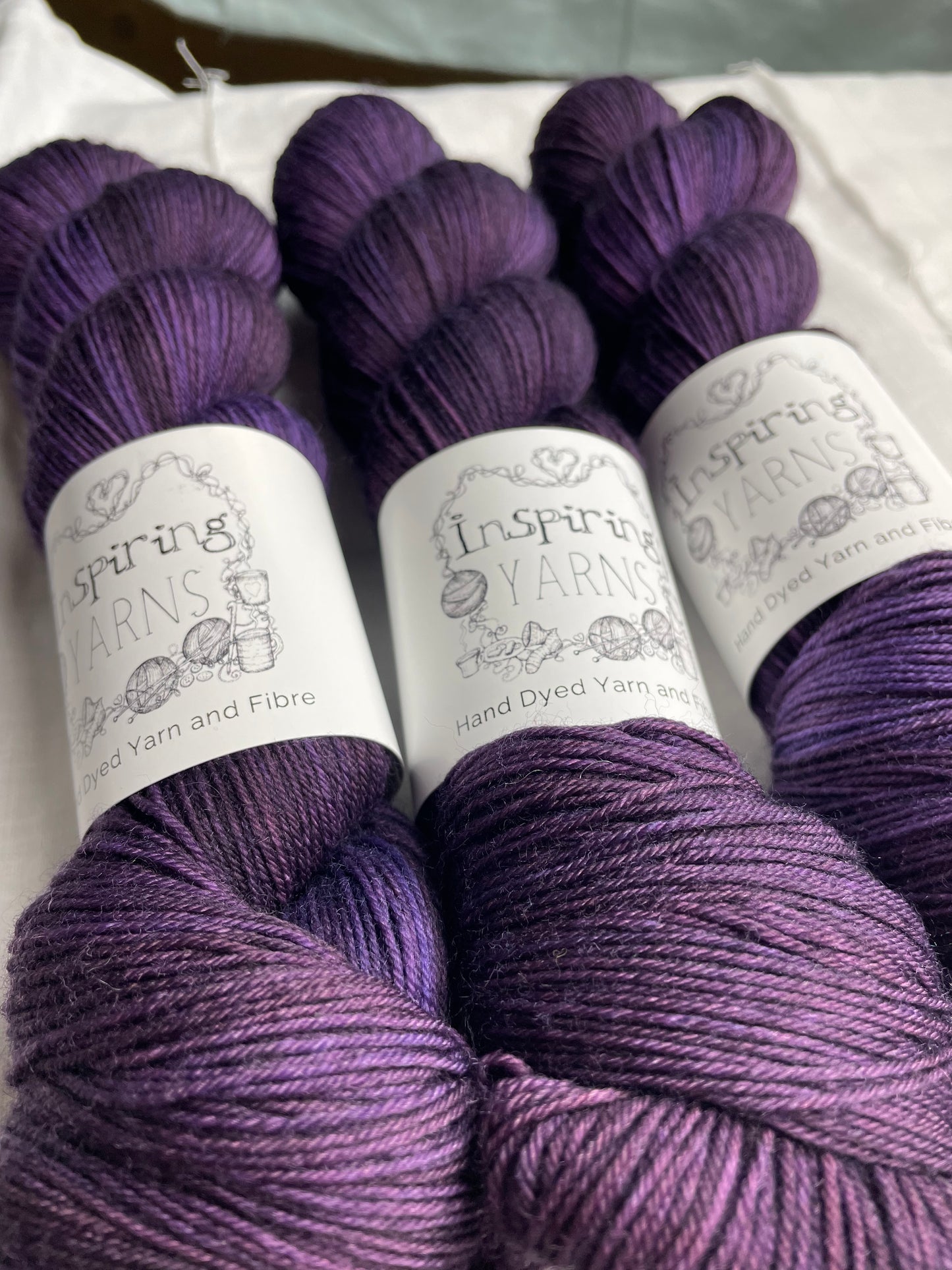 Three skeins of merino smooth sock yarn in a tonal dark purple colour labelled Inspiring Yarns on a white linen background.