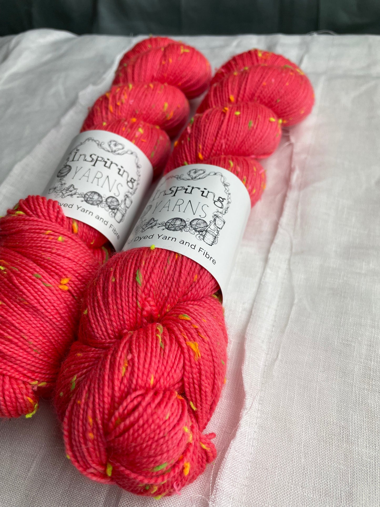 Close up of two skeins of tonal dark neon coral coloured hand dyed Disco Donegal yarn branded Inspiring Yarns on a white linen background. The yarn has multi coloured neon neps.