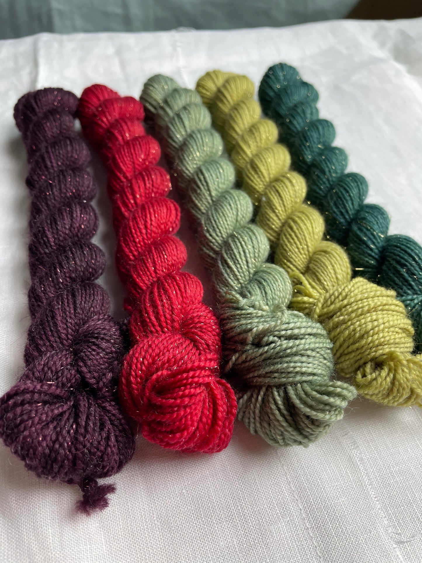 A set of five hand dyed yarn mini skeins on Sparkle Sock Yarn in red and green tones labelled Inspiring Yarns. The background is white linen and some Christmas lights with pinecones, twigs and stars.