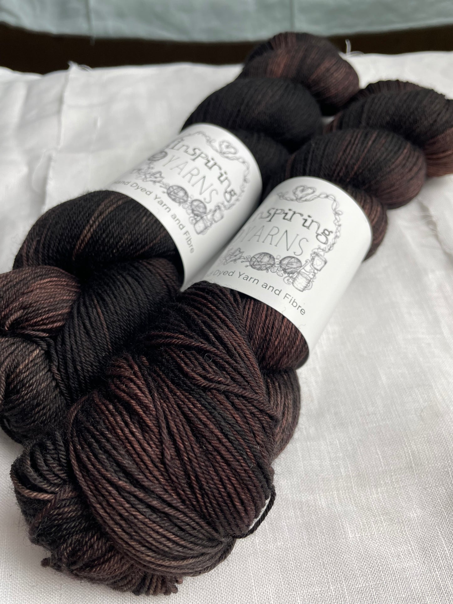 Two skeins of variegated dark brown black Merino Smooth Sock yarn labelled Inspiring Yarns on a white linen background