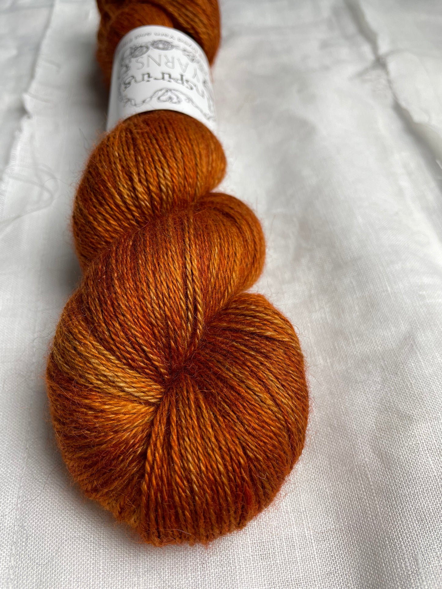 A skein of burnt orange mist 4ply yarn labelled Inspiring Yarns on a white linen background.