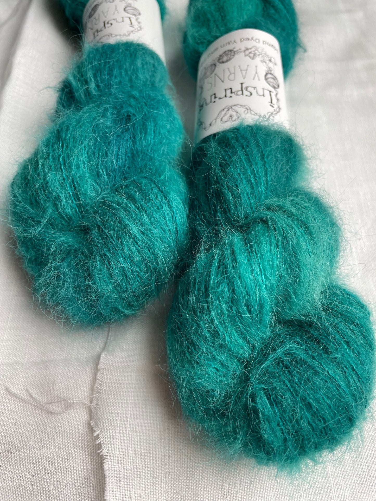 Close up of two skeins of fluffy brushed suri silk lace yarn hand dyed in a tonal jade green colourway and labelled Inspiring Yarns on a white linen background.
