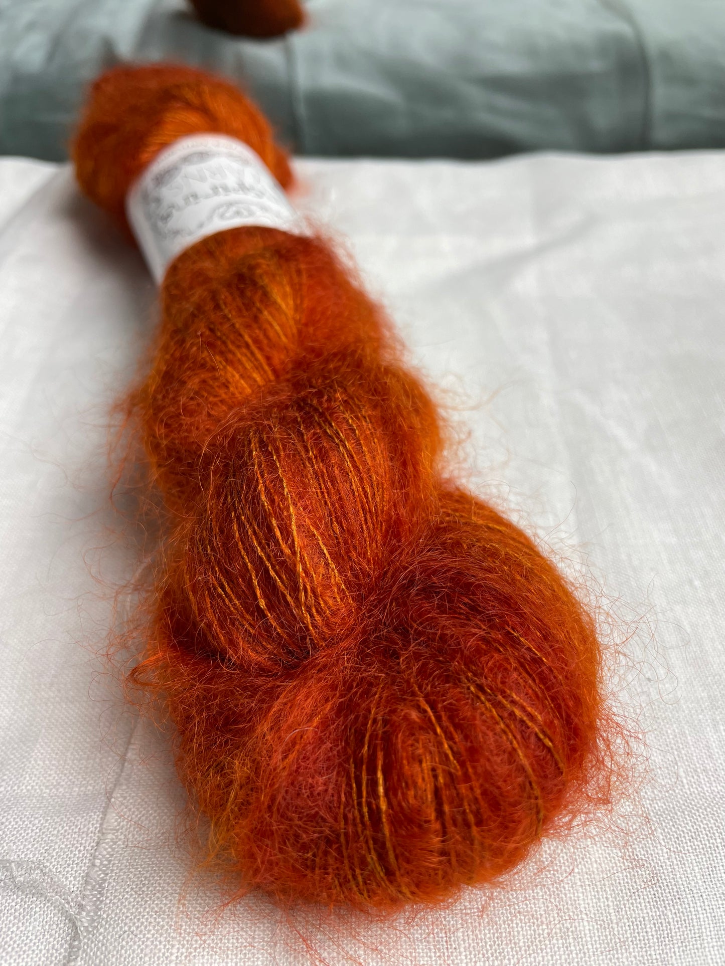 Close up of A skein of burnt orange kid mohair silk lace yarn labelled Inspiring Yarns on a white linen background.