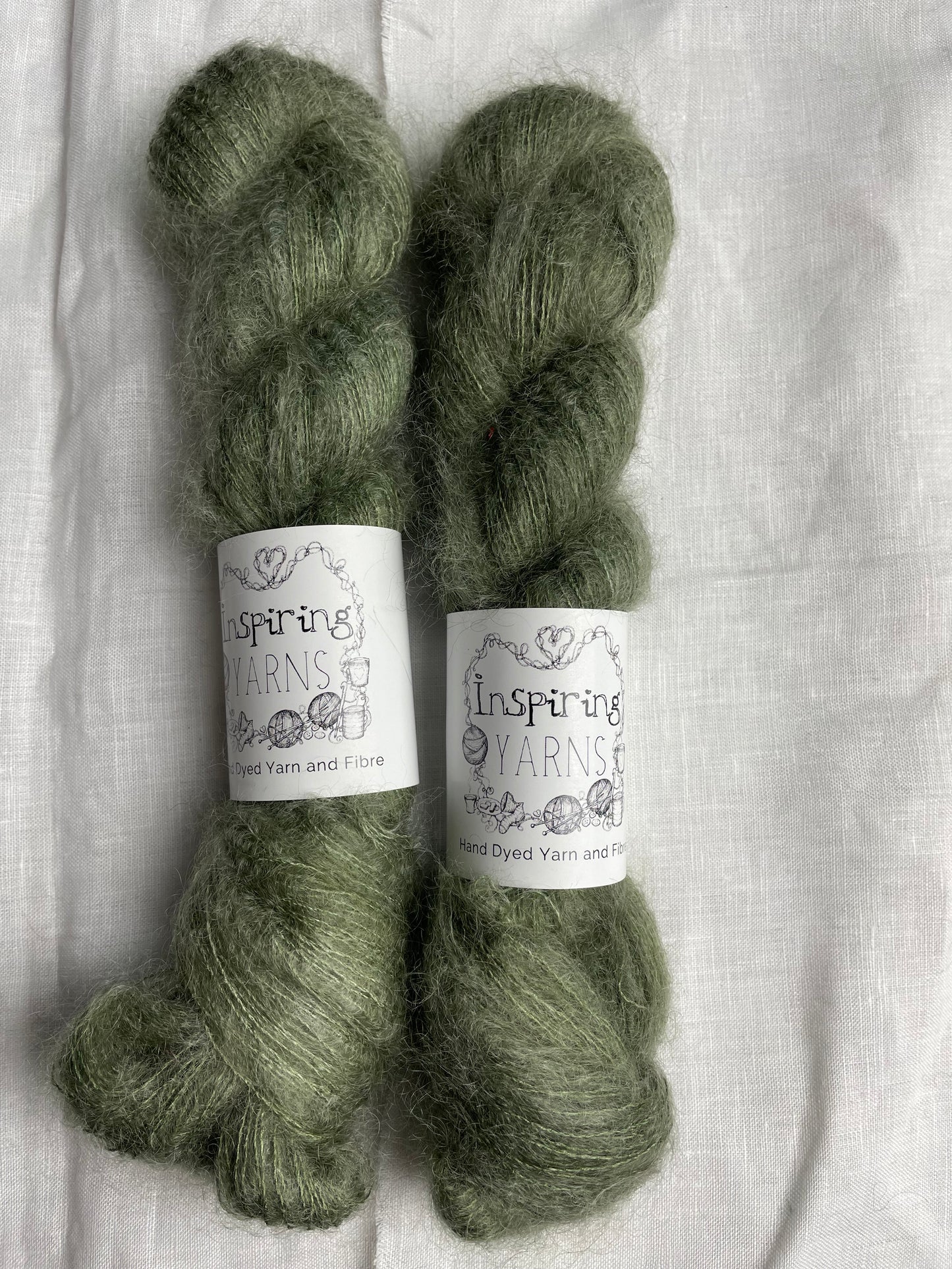 Two skeins of light grey green mohair silk lace yarn labelled Inspiring Yarns on a white linen background.