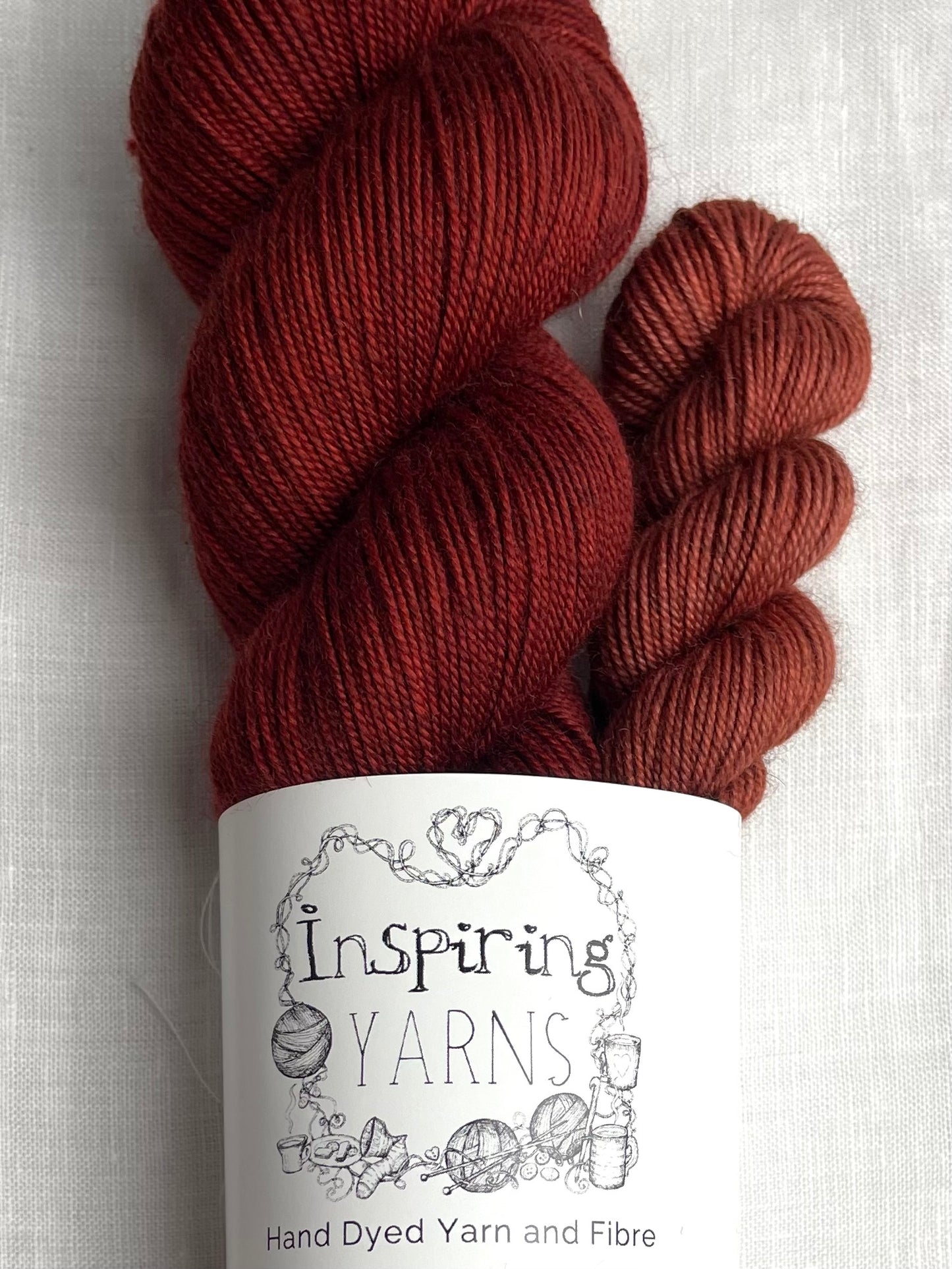 Close up of a sock set made of a skein of dark tonal rusty red hand dyed yarn with a contrasting deep terraccotta mini skein, labelled Inspiring Yarns and laid on a white linen cloth.