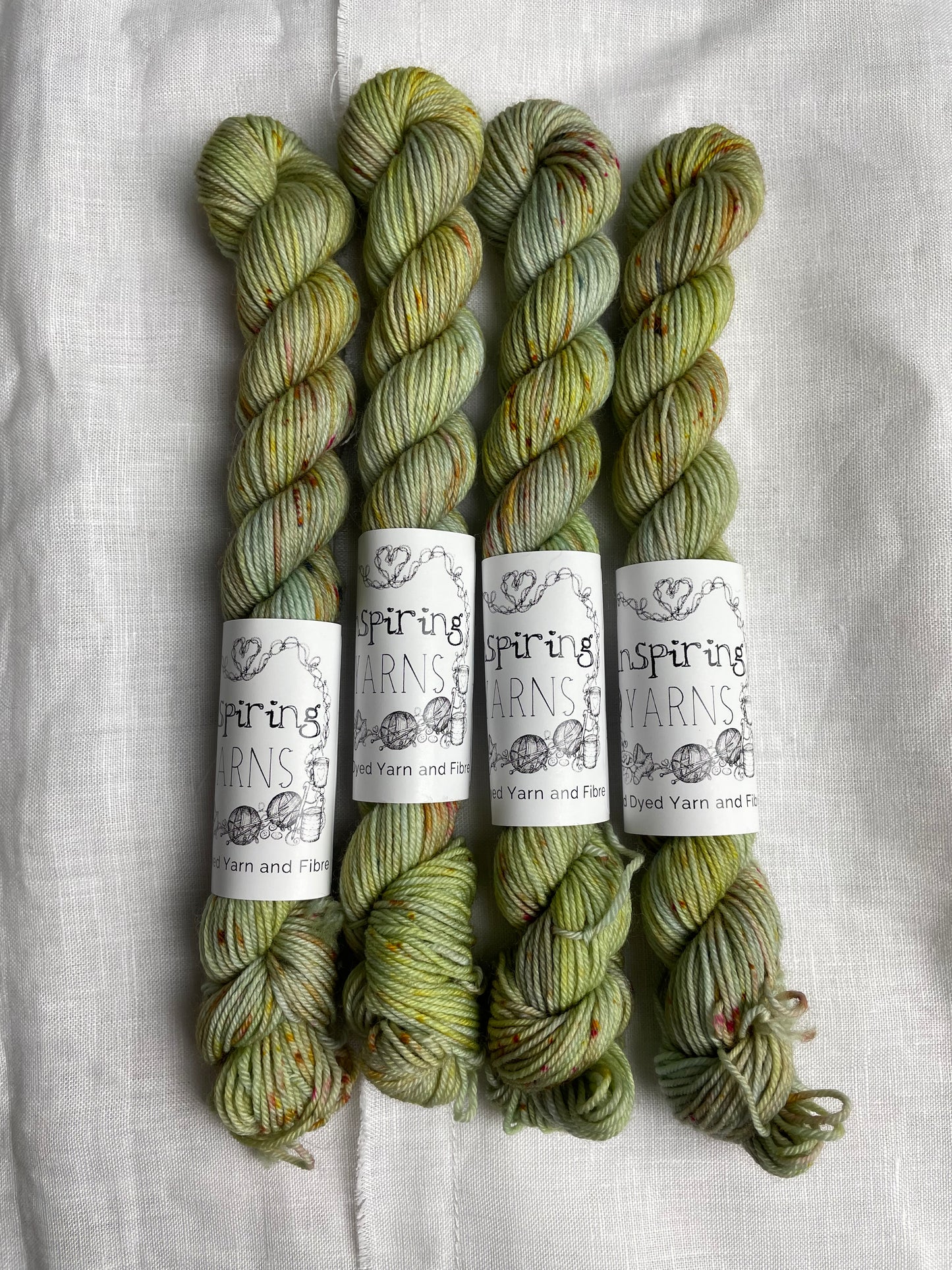 Four mini skeins of Faded Glory, a pale green with speckles coloured Merino Smooth Sock, labelled Inspiring Yarns on a white linen background.