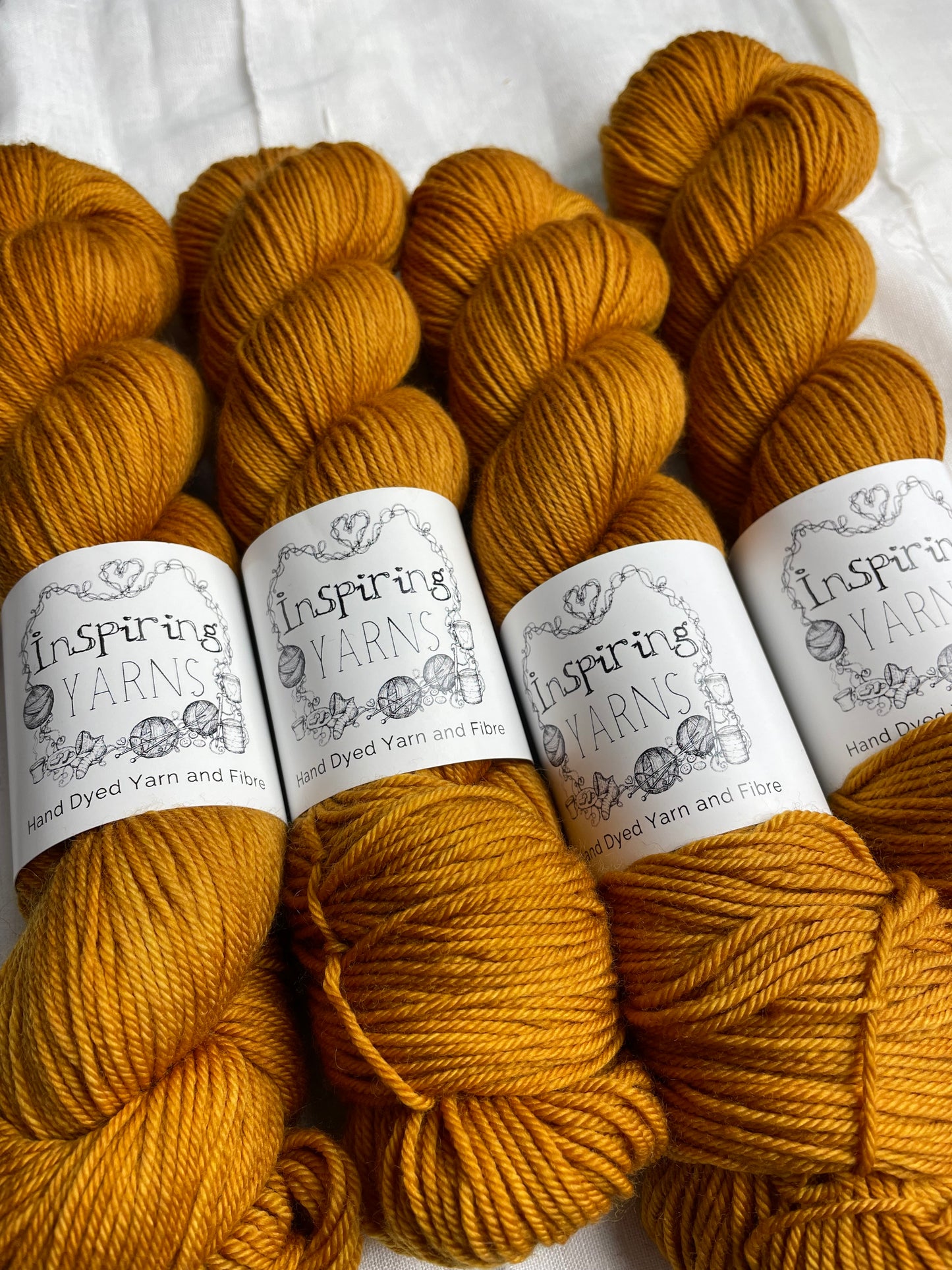 Close up of four skeins of mustard Merino Smooth DK yarn labelled Inspiring Yarns on a white linen background.