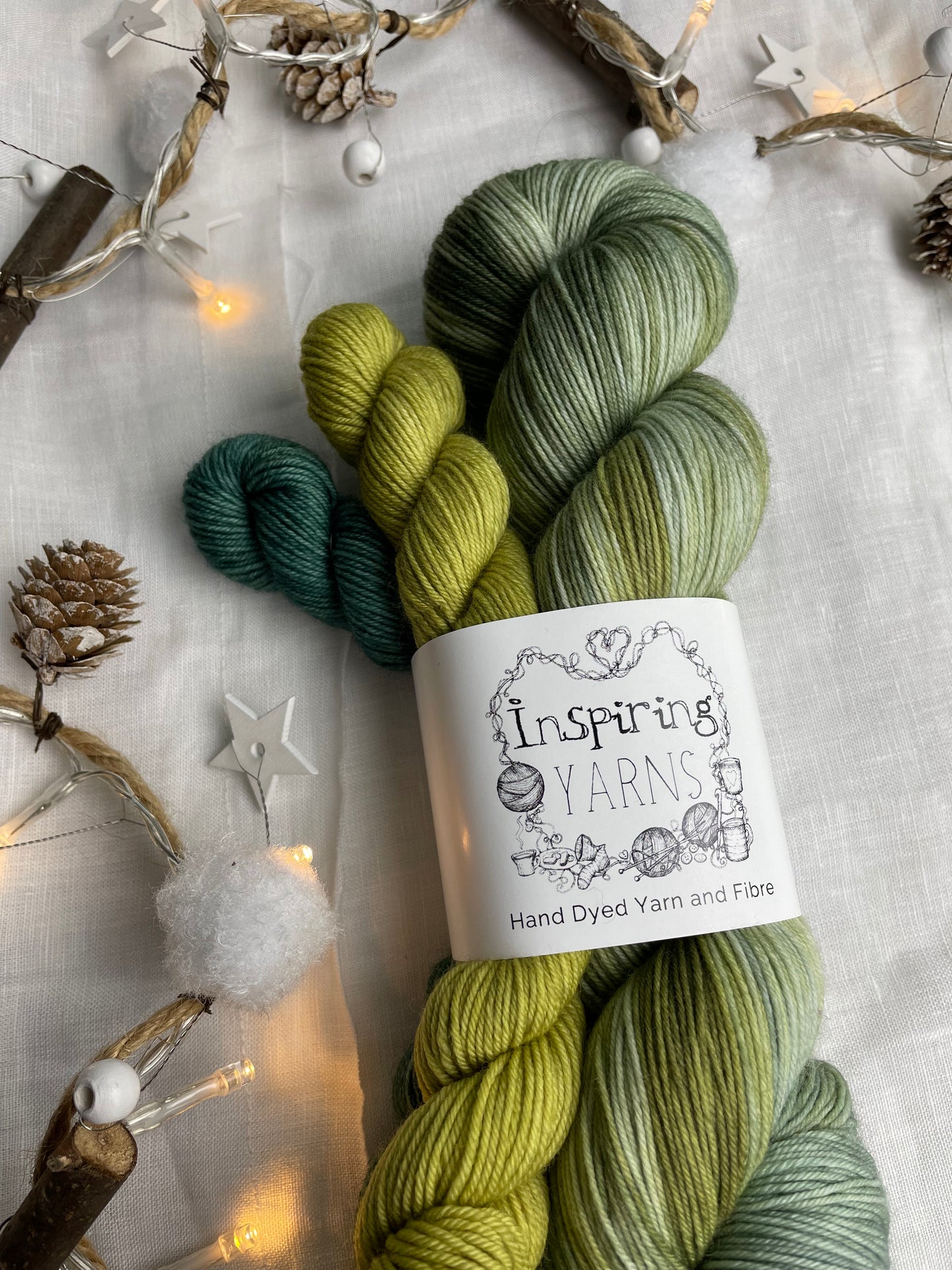 A hand dyed yarn sock set featuring large skein in green tones with a teal and a lime green mini skein labelled Inspiring Yarns. The background is white linen and some Christmas lights with pinecones, twigs and stars.