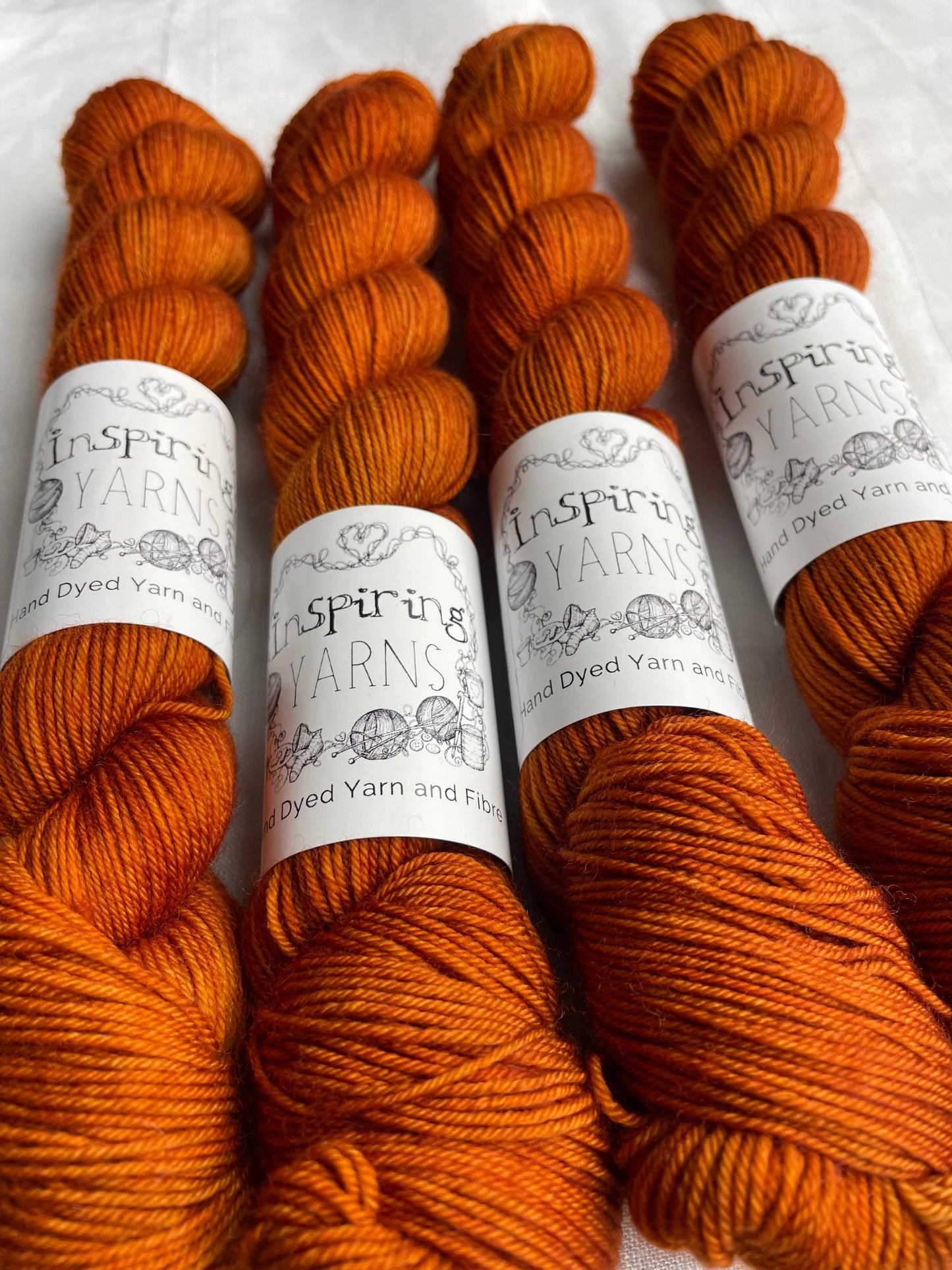 Four tonal burnt orange coloured skeins of hand dyed Merino Smooth Sock yarn branded Inspiring Yarns on white linen fabric.