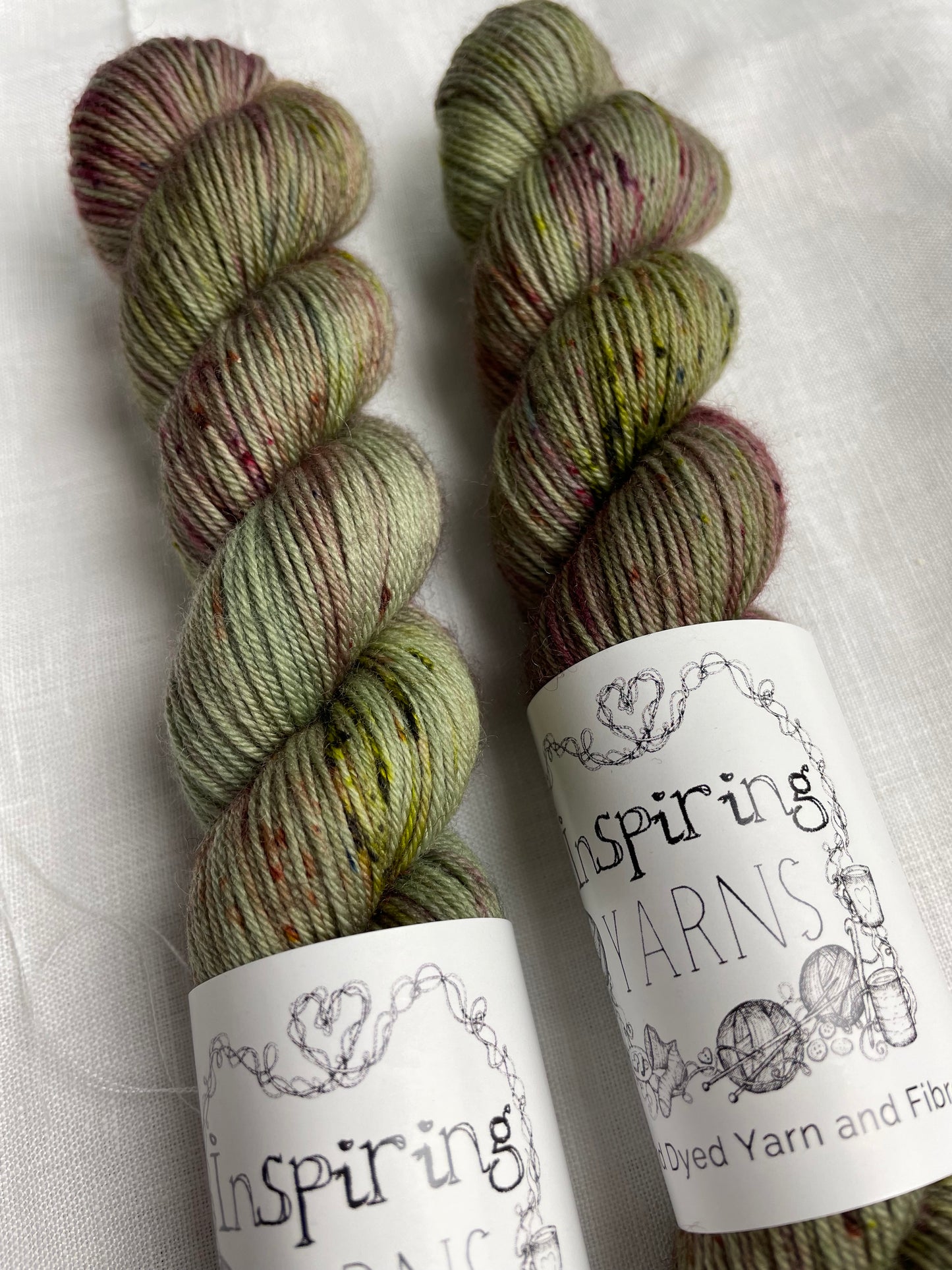 Two skeins of Cassidy, a sage green with speckles coloured Merino Smooth Sock, labelled Inspiring Yarns on a white linen background.