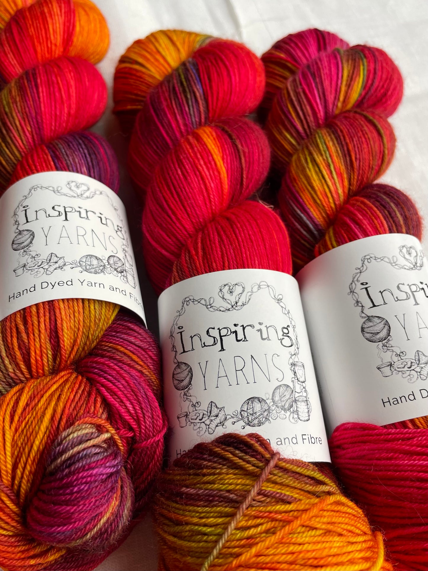Close up of four variegated red and multi coloured skeins of Merino Smooth Sock yarn branded Inspiring Yarns on a white linen fabric background.