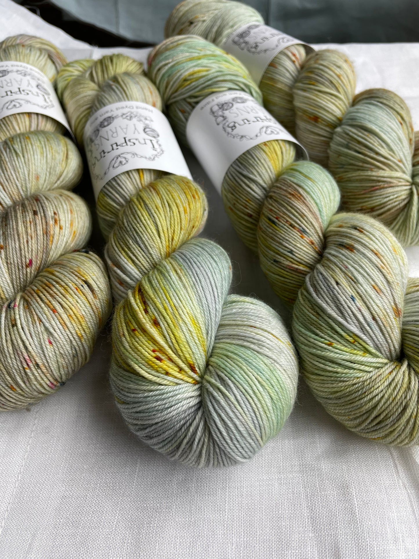 Close up of four lightly speckled, variegated grey and green coloured skeins of Merino Smooth Sock yarn branded Inspiring Yarns on a white linen fabric background.