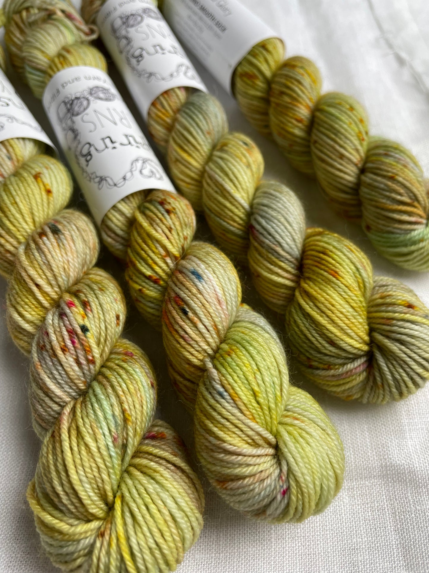 Close up of four yellow based speckled mini skeins of Merino Smooth Sock yarn branded Inspiring Yarns on a white linen fabric background.
