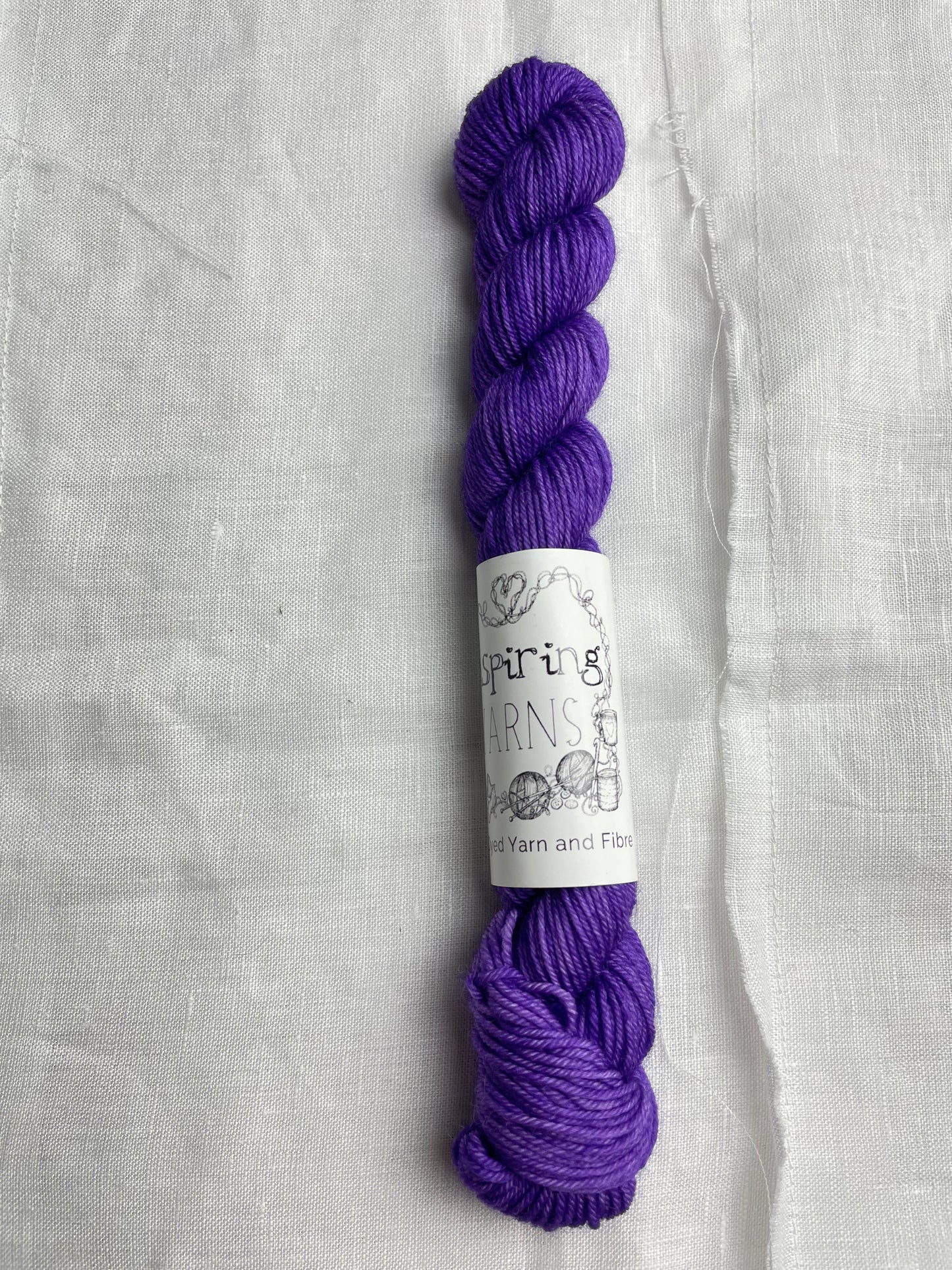 A bright purple coloured mini skein of Merino Smooth Sock yarn branded Inspiring Yarns on a white linen fabric background.