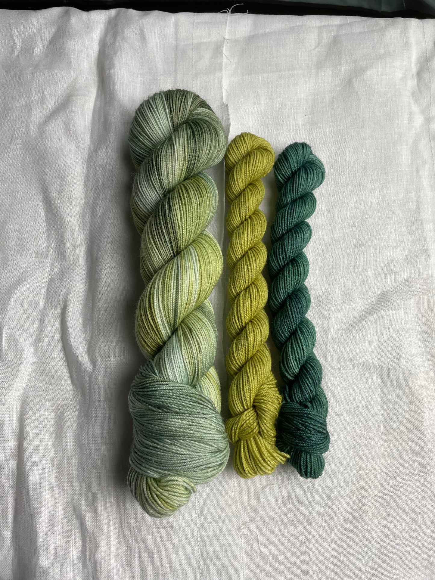 A hand dyed yarn sock set featuring large skein in green tones with a teal and a lime green mini skein labelled Inspiring Yarns. The background is white linen and some Christmas lights with pinecones, twigs and stars.
