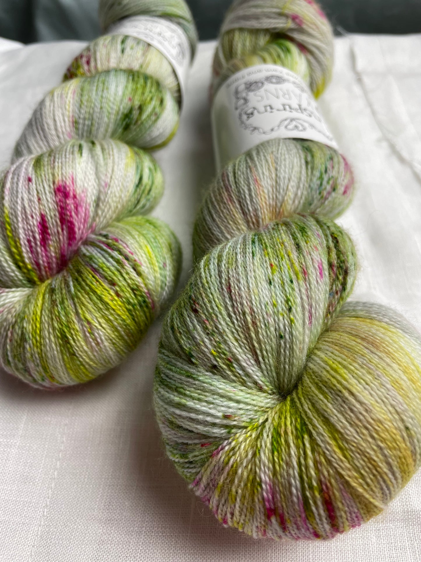Close up of two skeins of pink and green speckled silver grey coloured hand dyed Merino Lace yarn branded Inspiring Yarns on a white linen cloth.