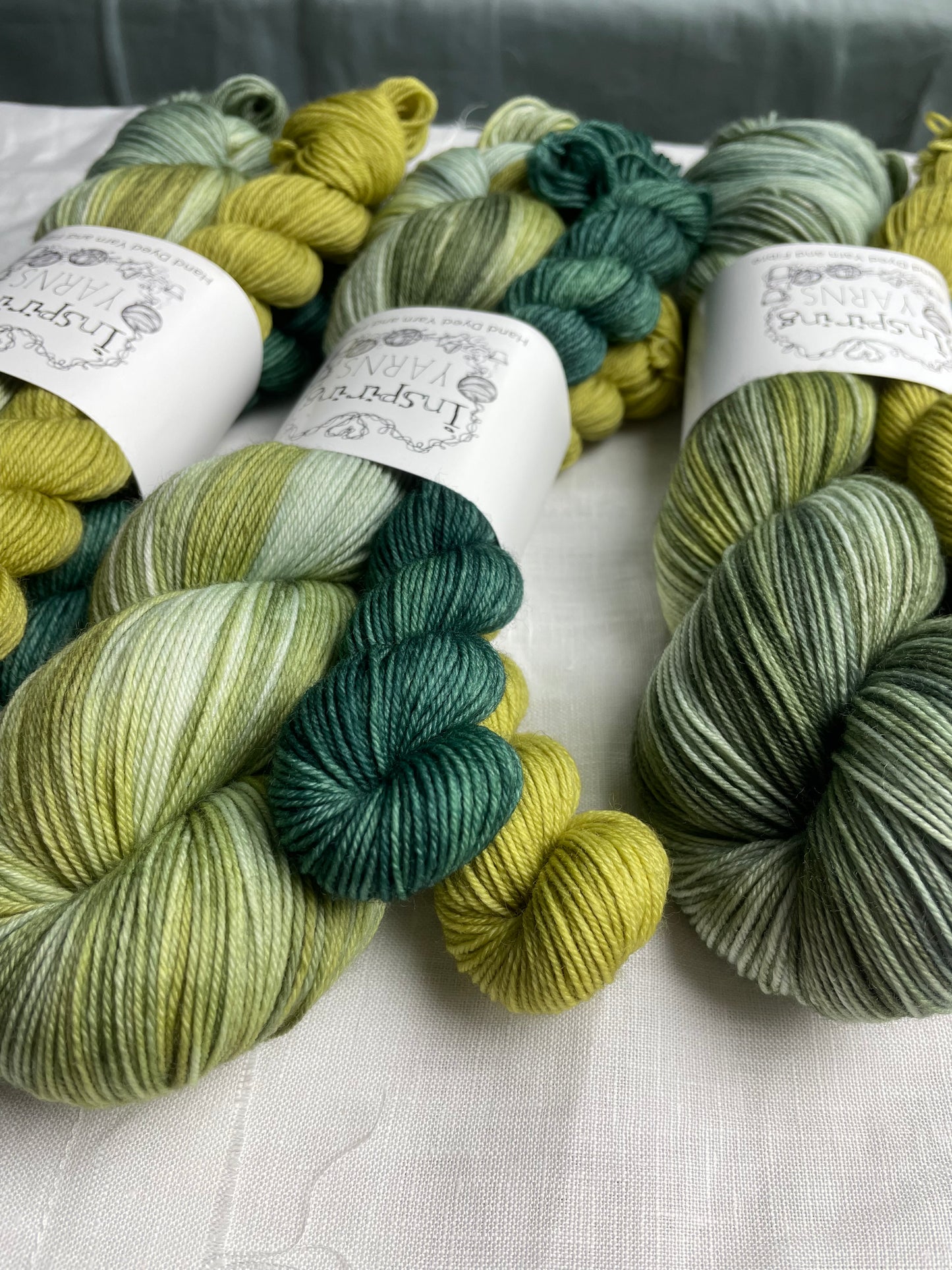 Three hand dyed yarn sock sets featuring large skein in green tones with a teal and a lime green mini skein labelled Inspiring Yarns. The background is white linen and some Christmas lights with pinecones, twigs and stars.