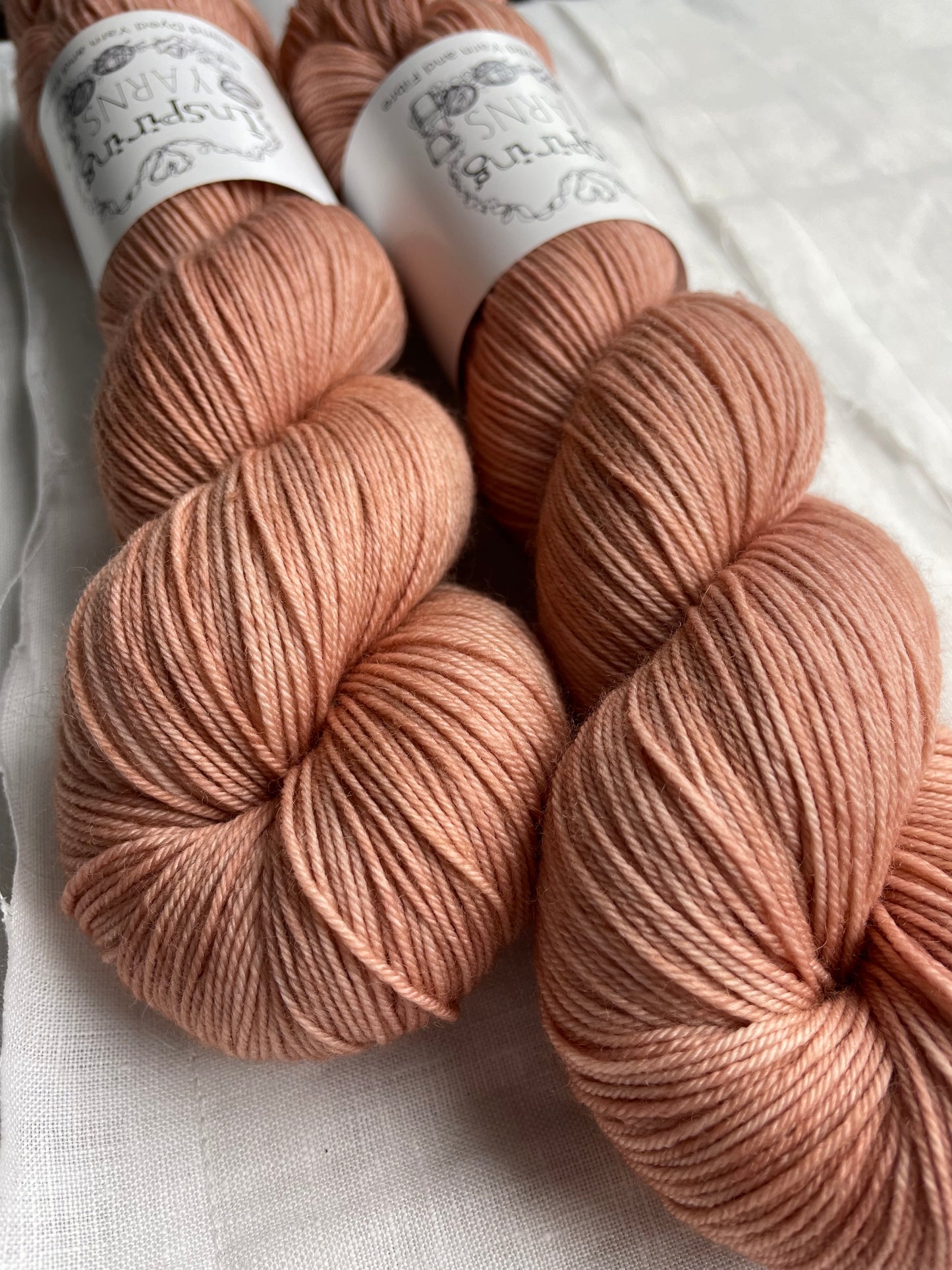 Close up of two dusky peach coloured skeins of Merino Smooth Sock yarn branded Inspiring Yarns on a white linen fabric background.