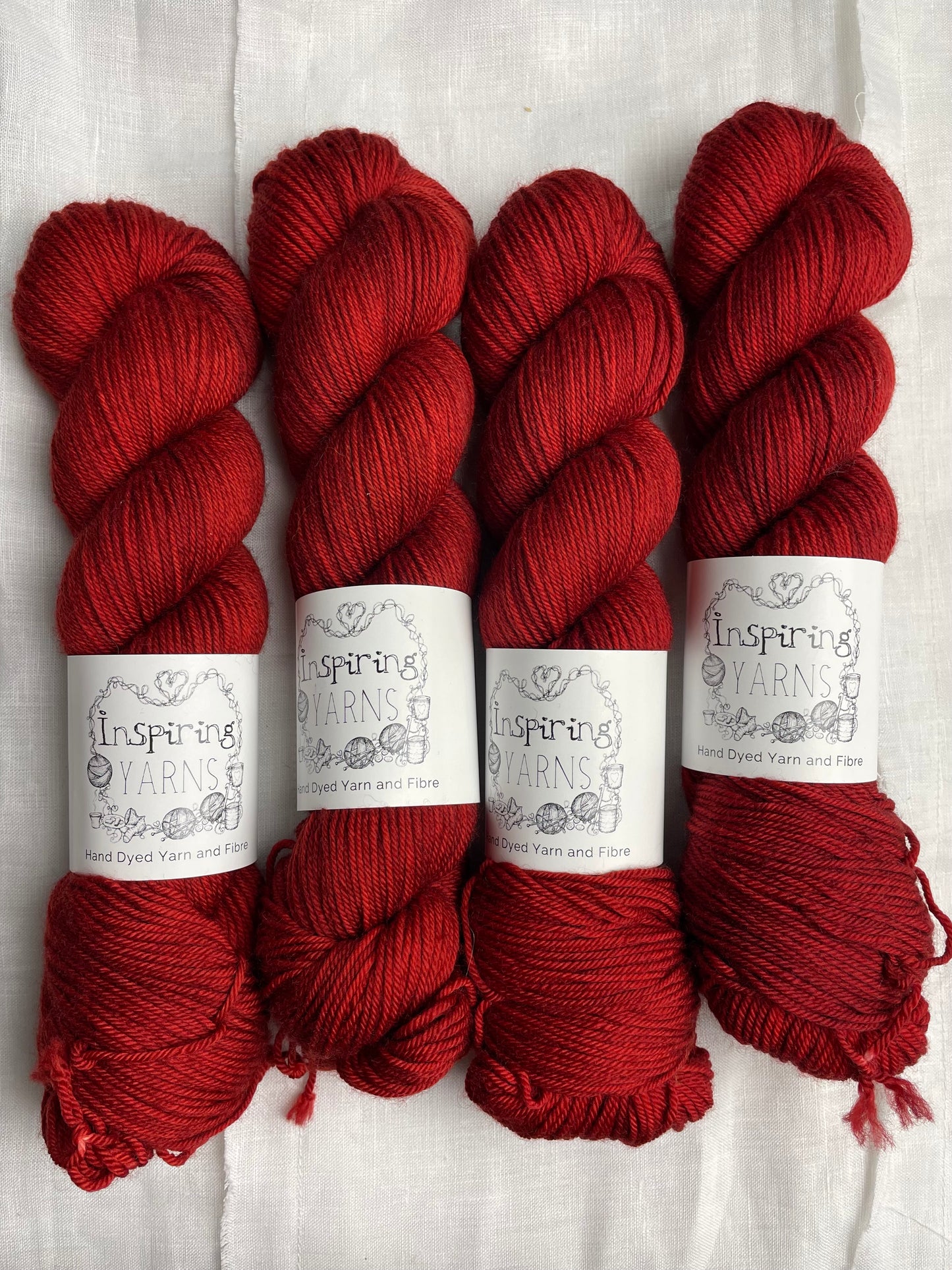 Four skeins of handyed Merino Smooth DK yarn in a rusty red colourway labelled Inspiring Yarns on a white linen background.