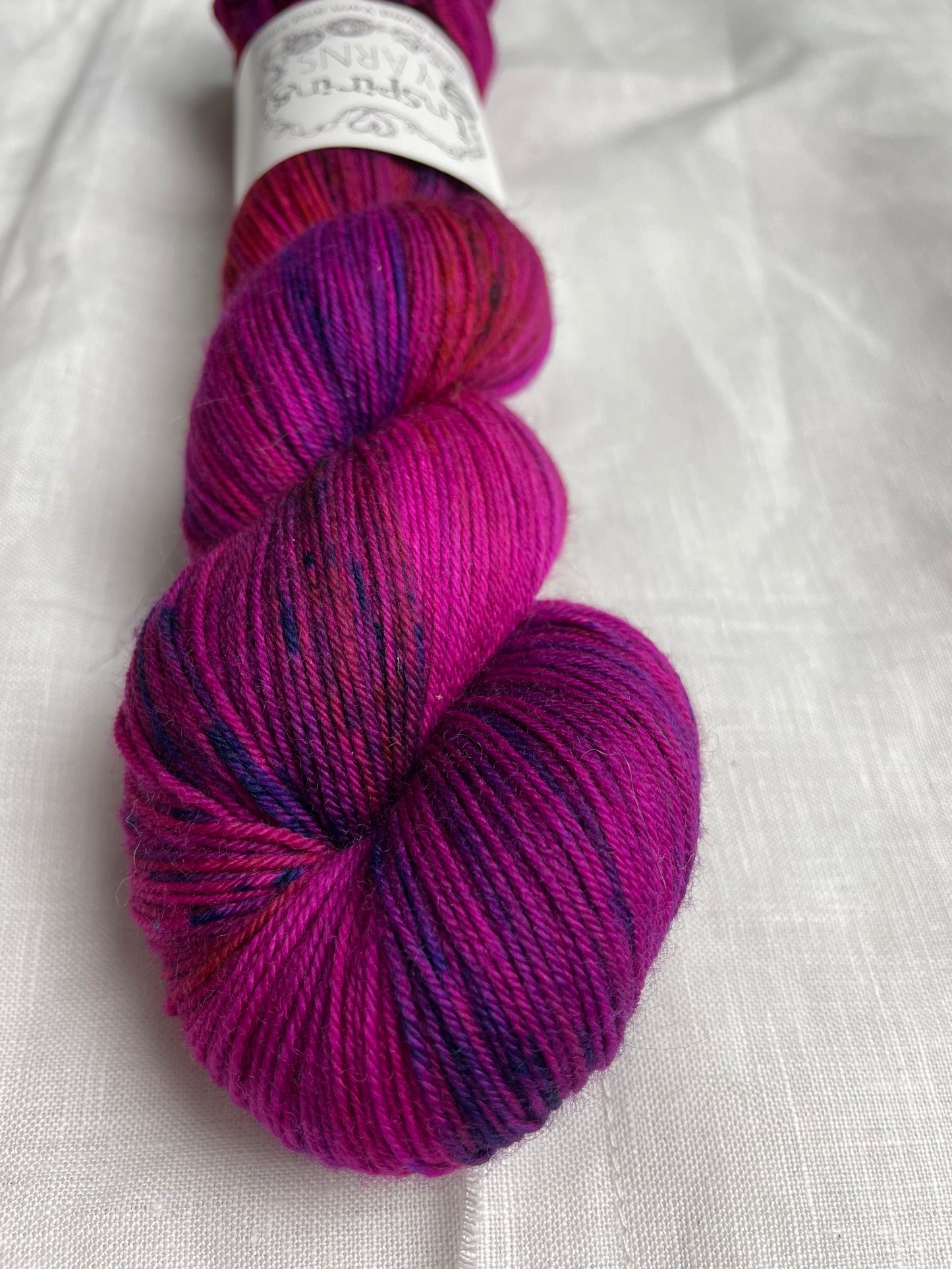 A skein of fuchsia pink speckled Merino Smooth Sock labelled InspiringYarns lies on a white linen background.