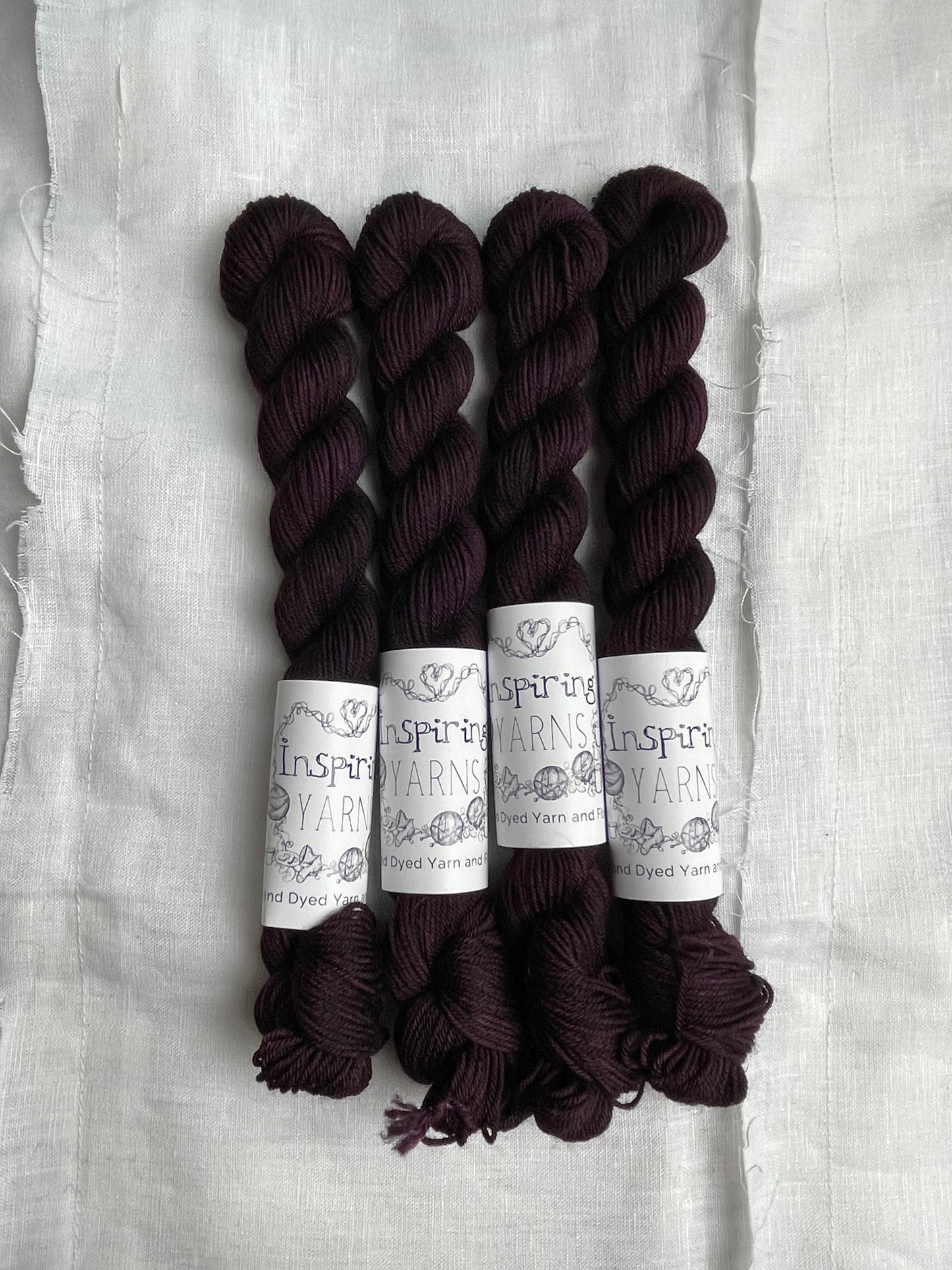 Four skeins of merino smooth sock yarn in a tonal dark burgundy colour labelled Inspiring Yarns on a white linen background.