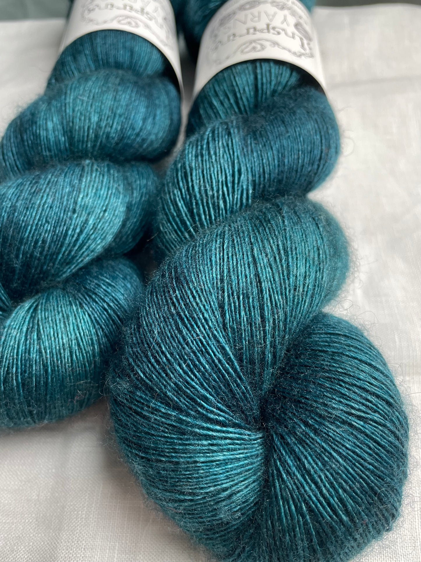 Close up of two tonal intense teal blue coloured skeins of hand dyed Yak Silk Singles branded Inspiring Yarns on a white linen background.