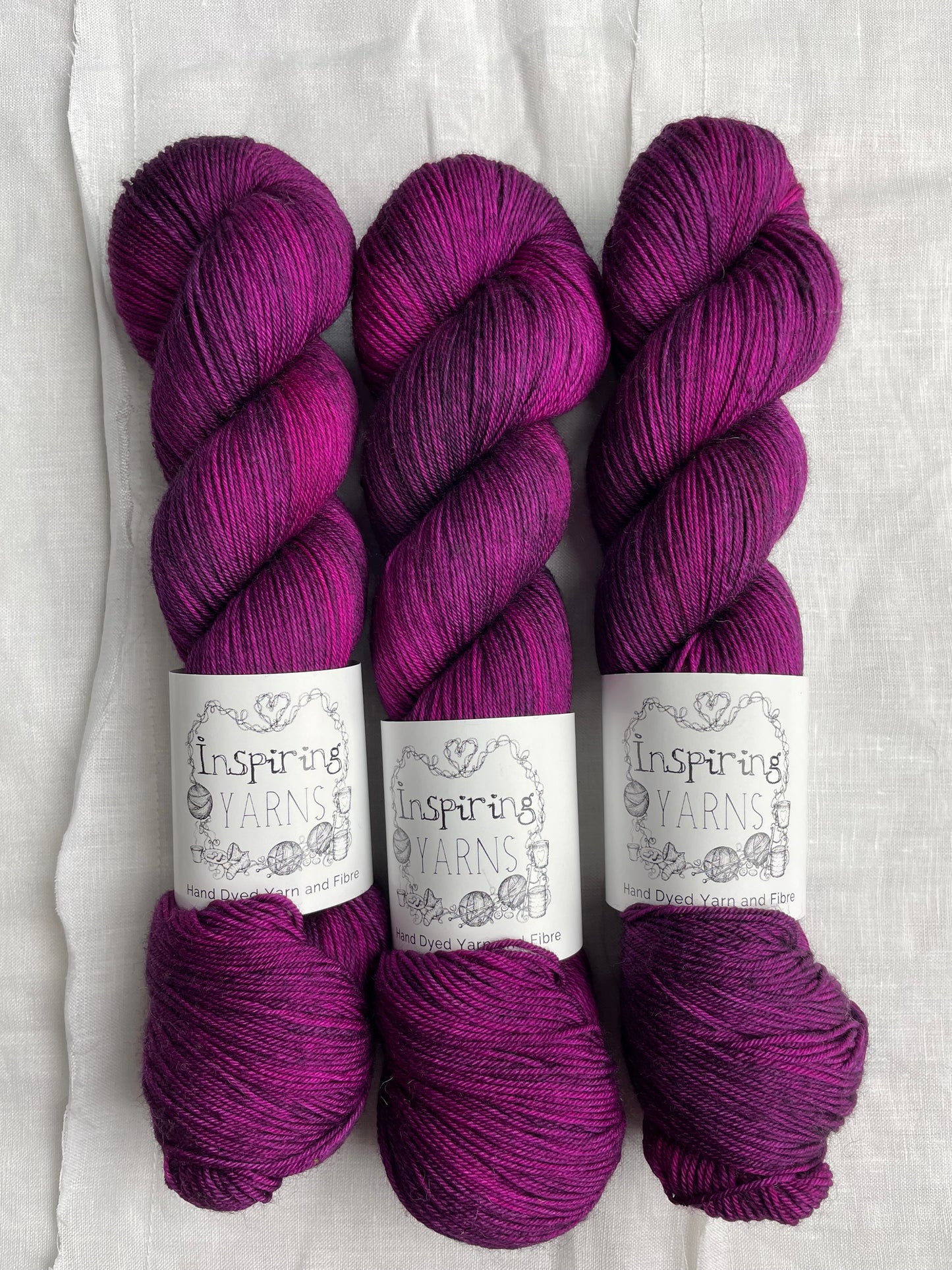 Three lightly variegated cerise pink and dark purple coloured skeins of Merino Smooth Sock yarn branded Inspiring Yarns on a white linen fabric background.