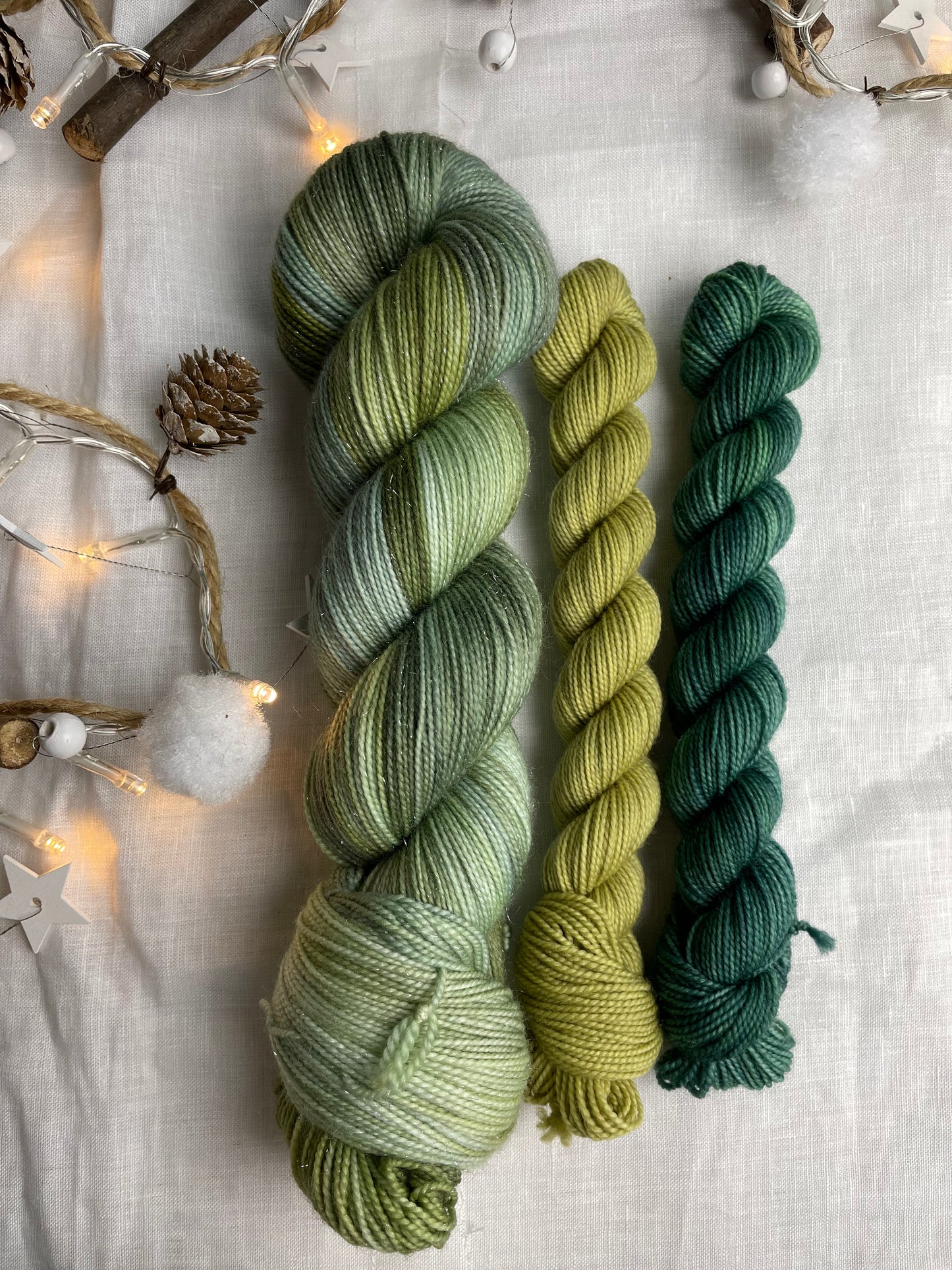 A hand dyed yarn sock set featuring large skein of Sparkle Sock Yarn in green tones with a teal and a lime green mini skein labelled Inspiring Yarns. The background is white linen and some Christmas lights with pinecones, twigs and stars.