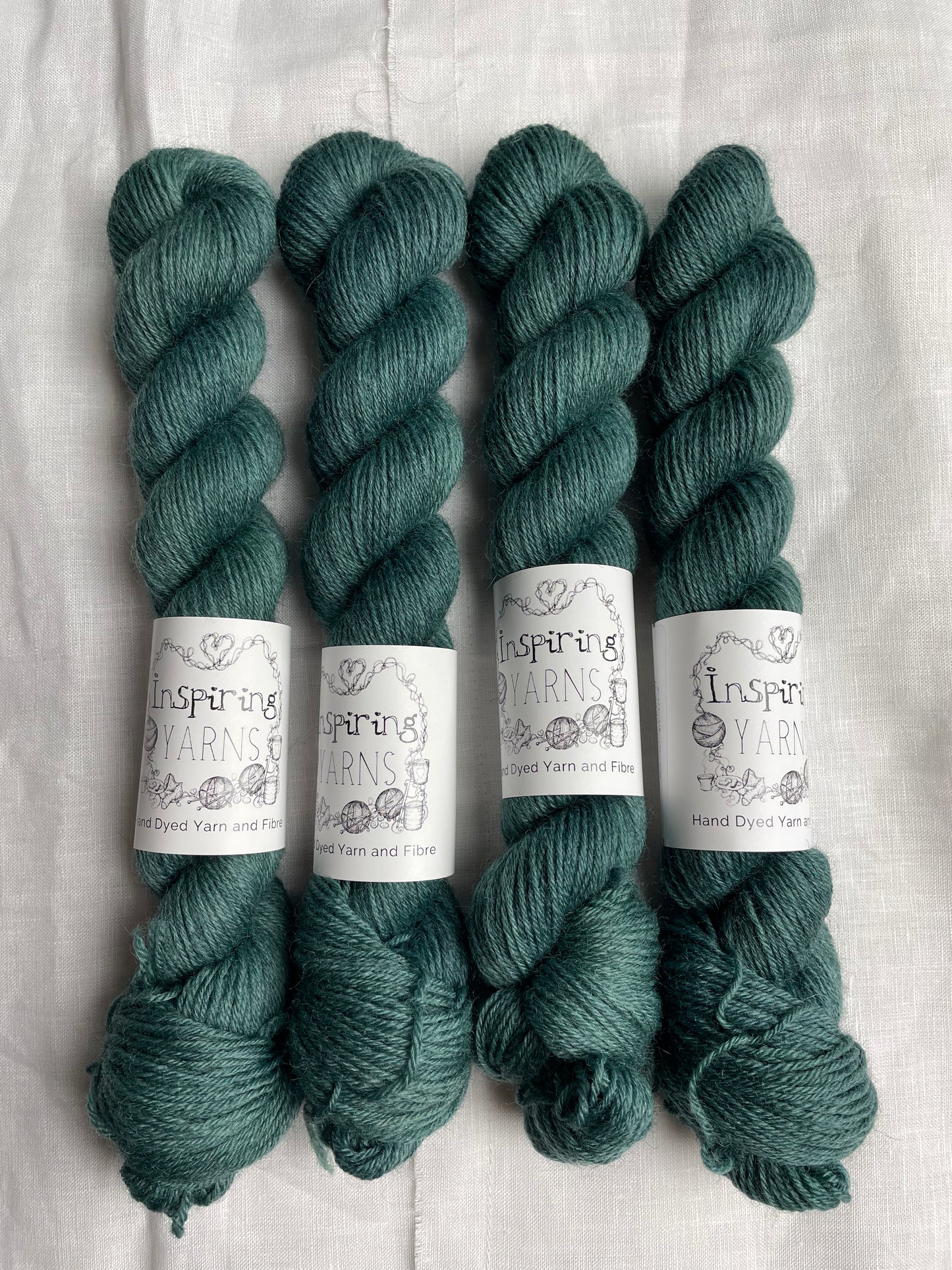 Four skeins of a tonal dark teal Highland 4Ply yarn branded Inspiring Yarns on a white linen background.