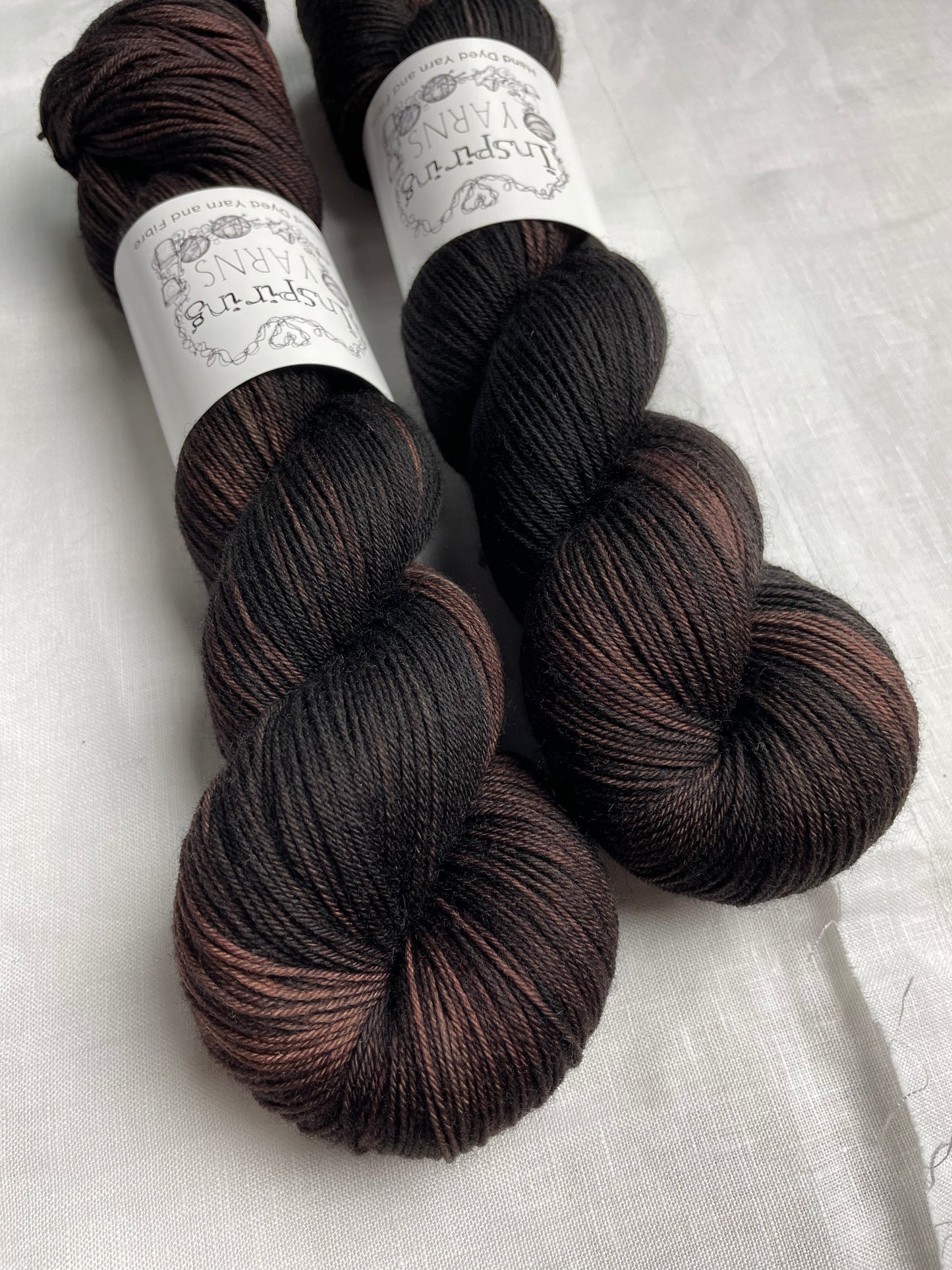 Two skeins of variegated dark brown black Merino Smooth Sock yarn labelled Inspiring Yarns on a white linen background