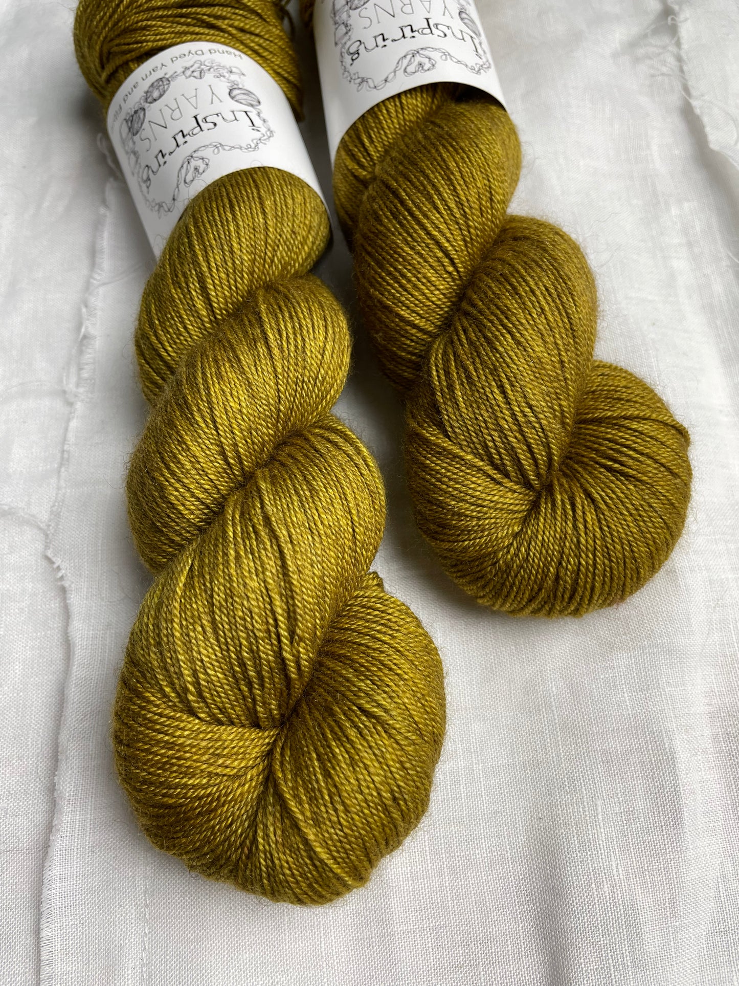 Close up of 2 skeins of ochre yellow Yak Silk 4ply yarn labelled Inspiring Yarns on a white linen background.