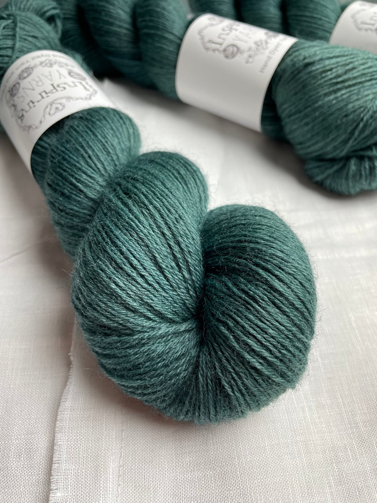 Three skeins of a tonal dark teal Highland 4Ply yarn branded Inspiring Yarns on a white linen background.