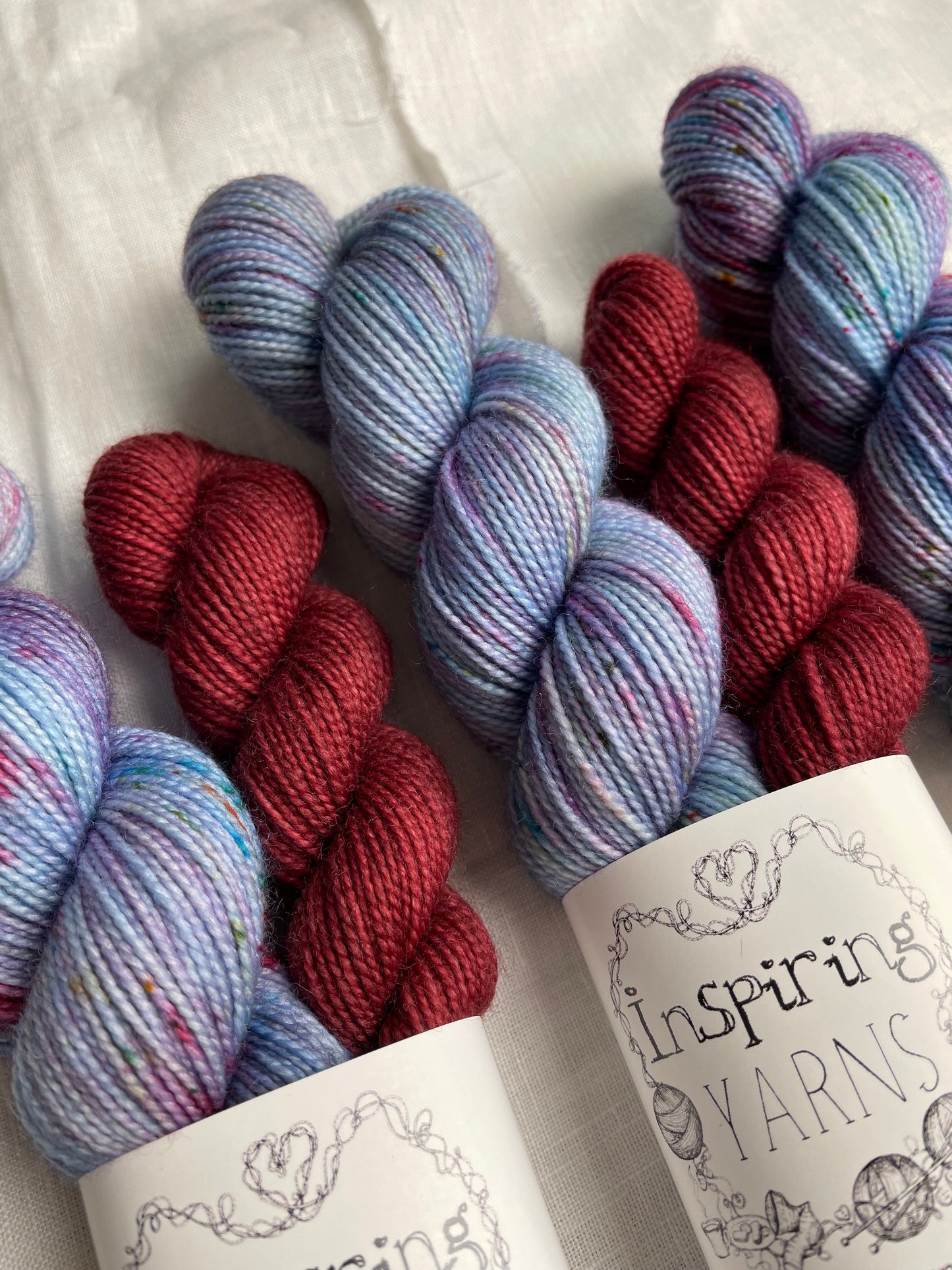Close up of three sock sets made of a skein of light blue speckled hand dyed yarn with a contrasting deep blood red mini skein, labelled Inspiring Yarns and laid on a white linen cloth.