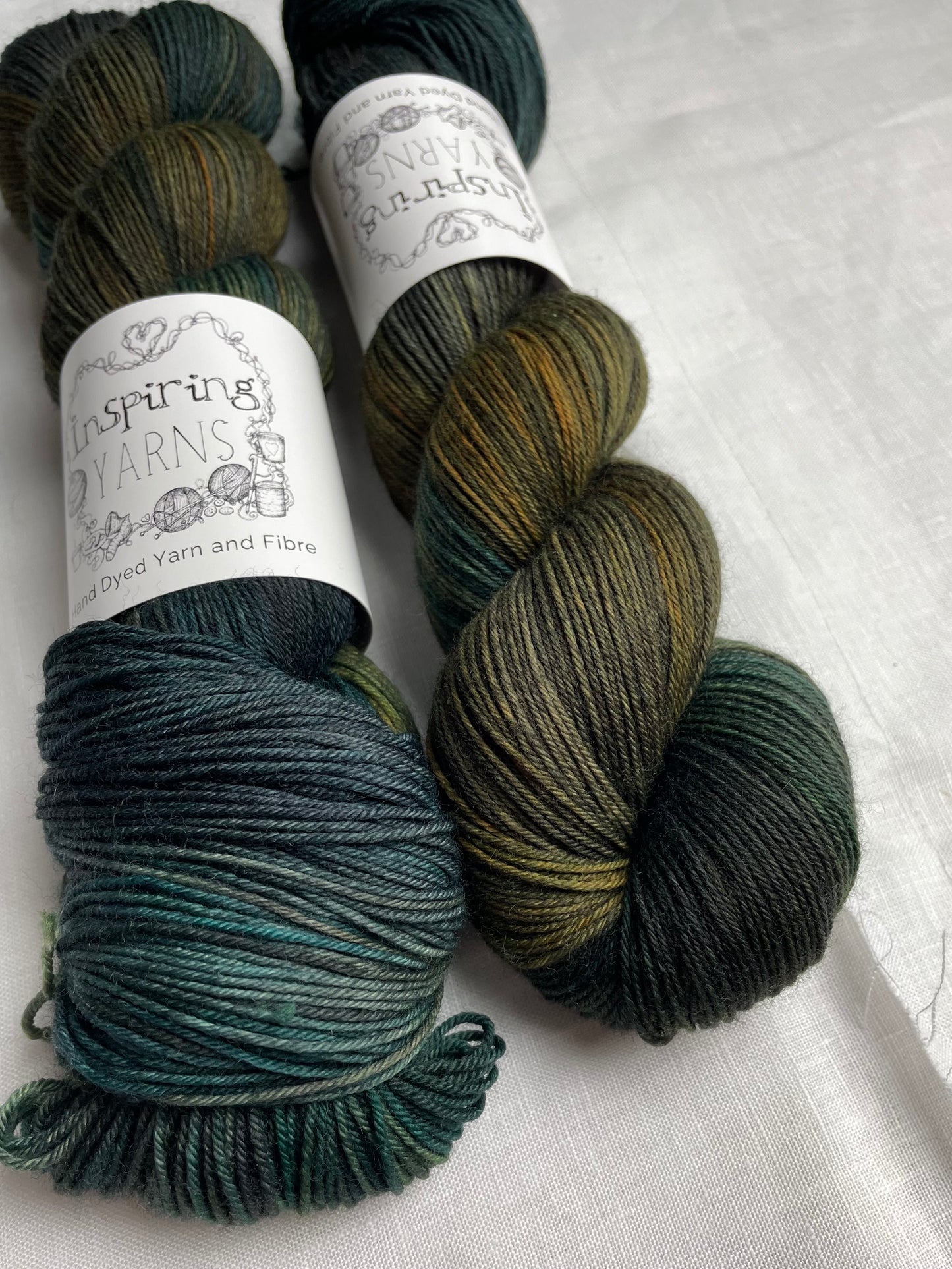 Two skeins of variegated black green gold and teal Merino Smooth Sock yarn labelled Inspiring Yarns on a white linen background