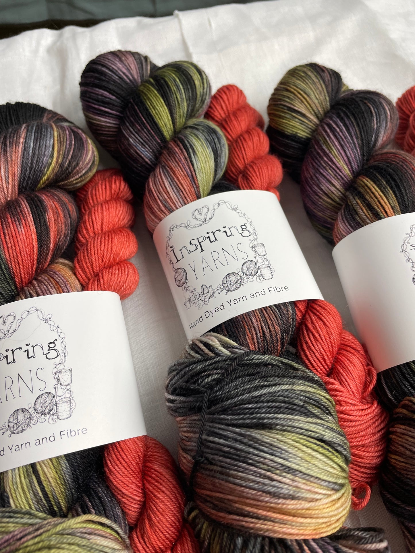 three sock sets made of a skein of black and multi coloured hand dyed yarn with a contrasting deep terraccotta mini skein, labelled Inspiring Yarns and laid on a white linen cloth.