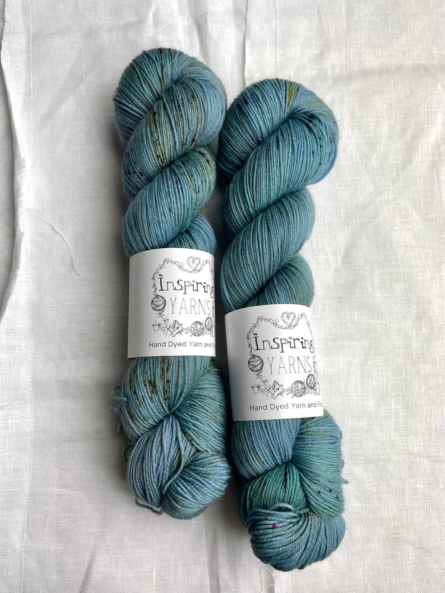 Two speckled soft teal coloured skeins of Merino Smooth Sock yarn branded Inspiring Yarns on a white linen fabric background.