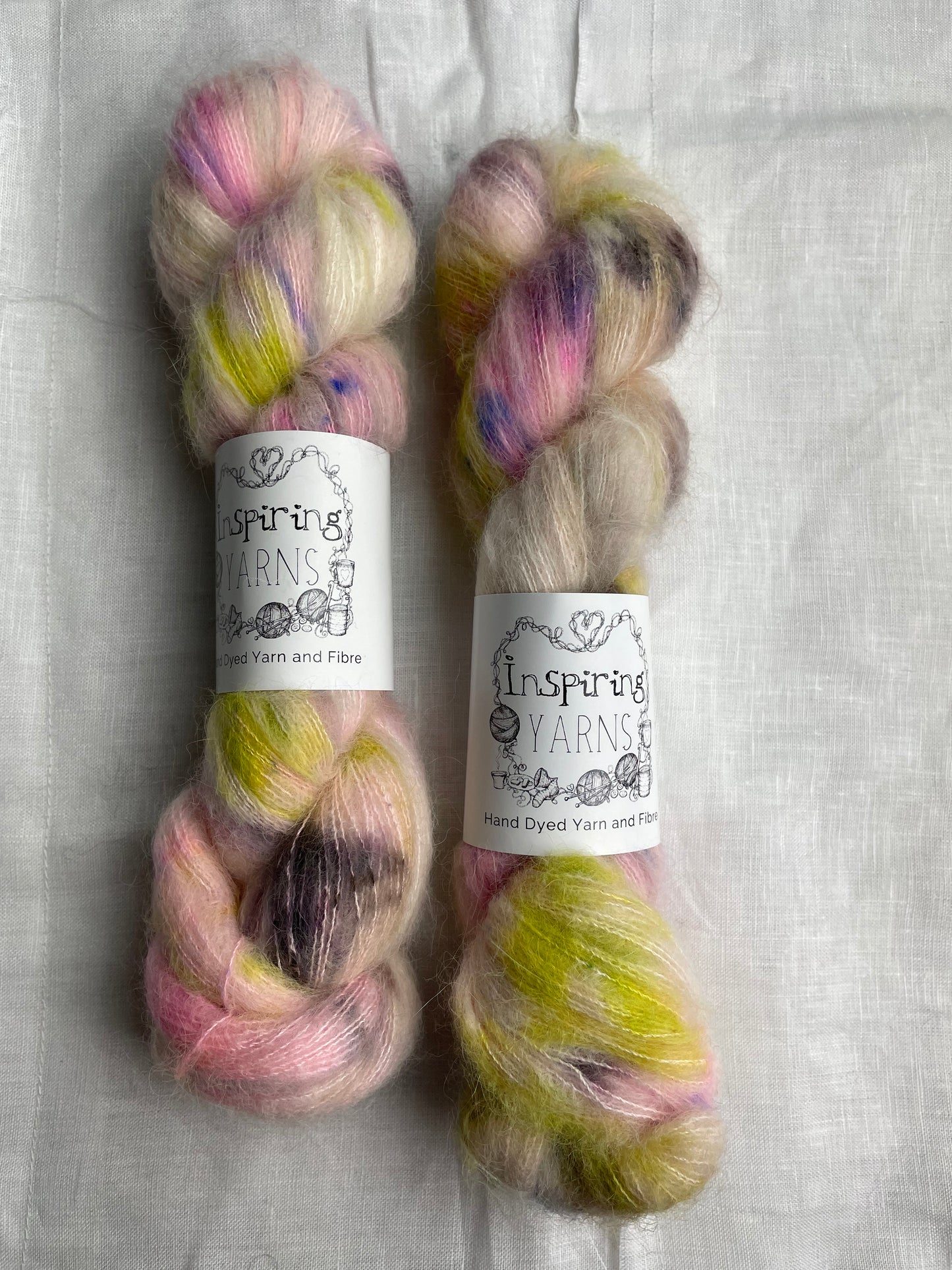 Two skeins of brightly speckled natural coloured hand dyed fluffy Alpaca Silk Lace yarn labelled Inspiring Yarns on a white linen background.