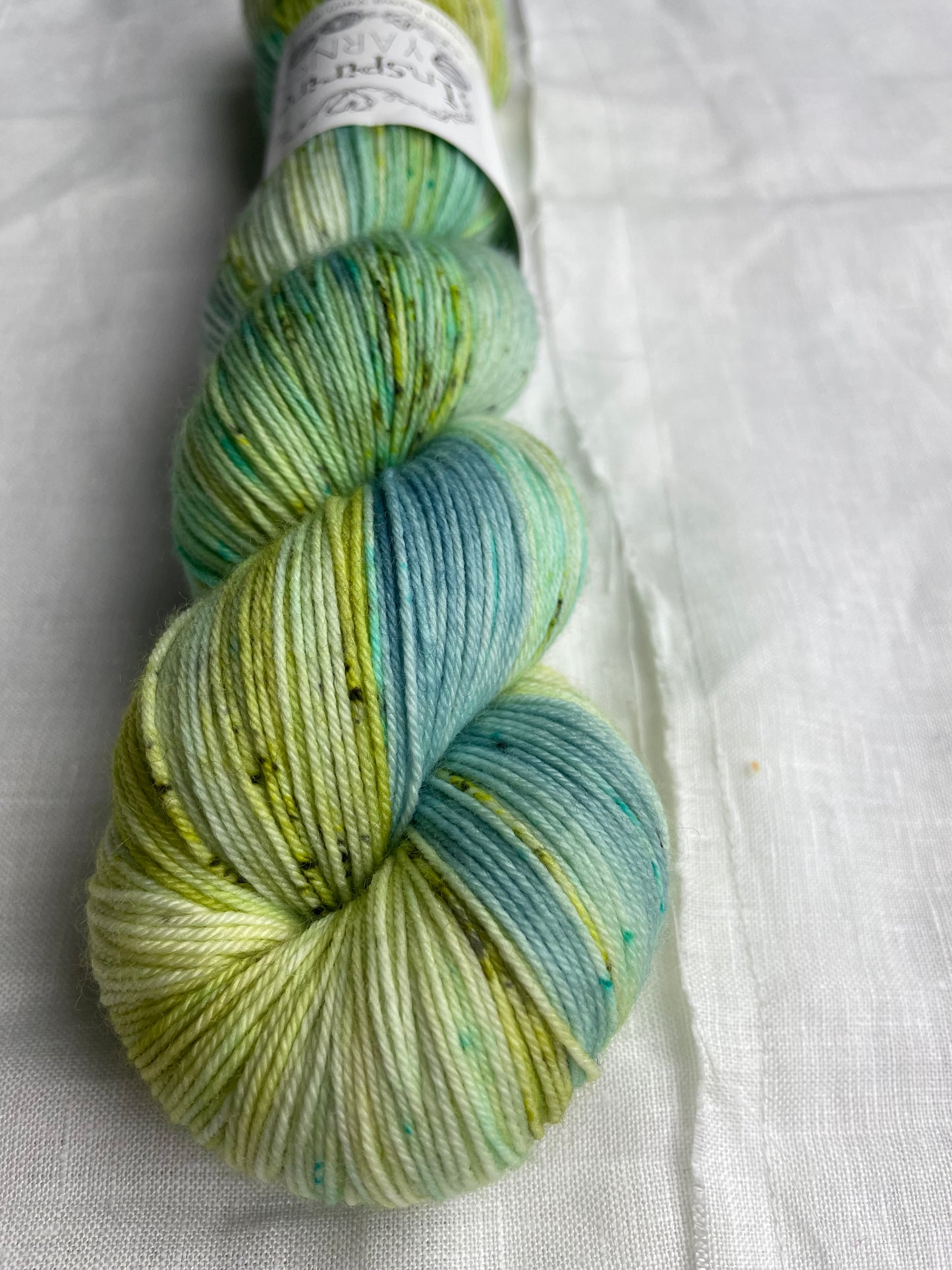 Close up of a skein of variegated blue, teal and acid yellow coloured Merino Smooth Sock yarn branded Inspiring Yarns on a white linen fabric background.