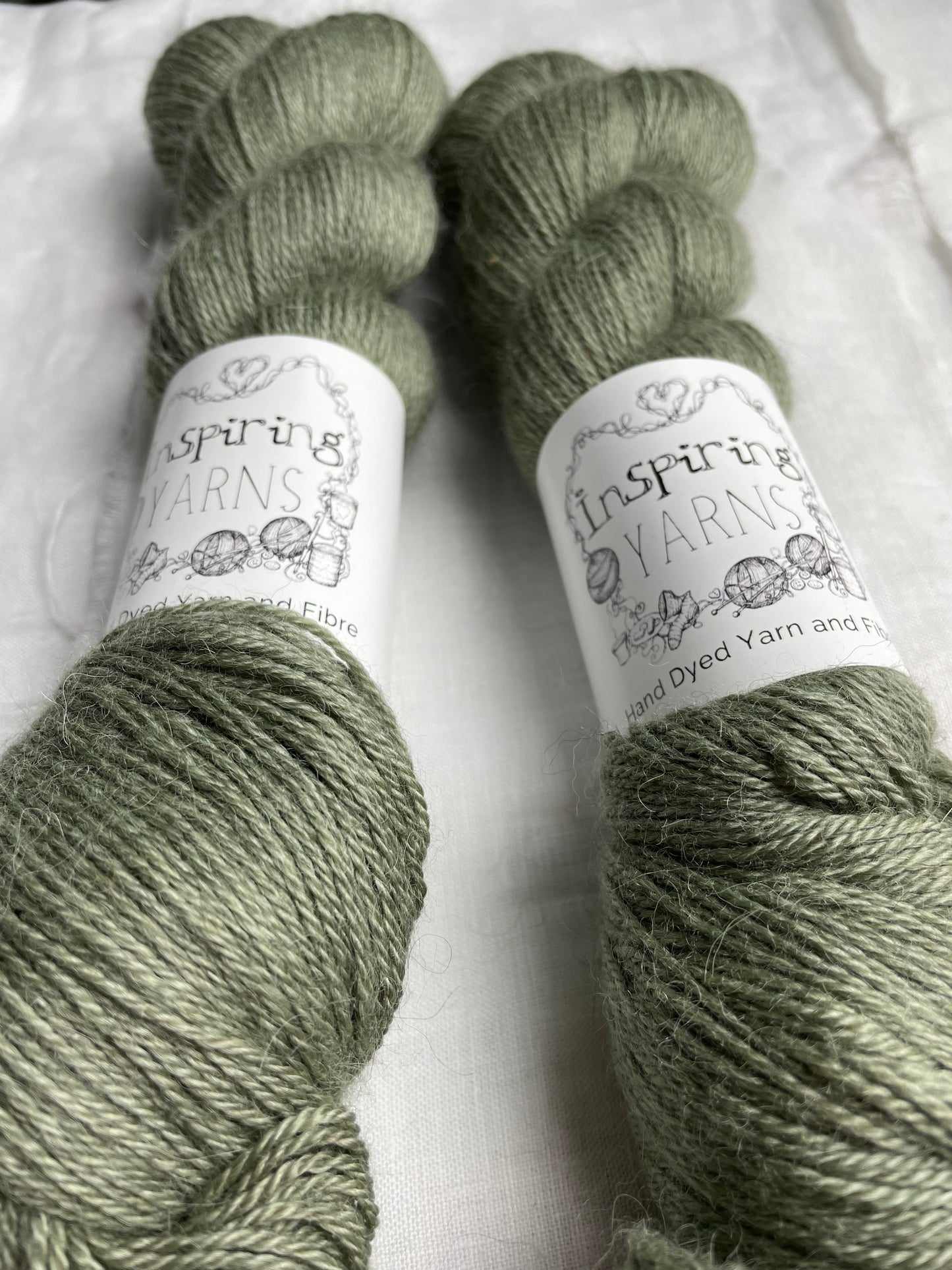 Close up of two skeins of pale green mist 4ply yarn labelled Inspiring Yarns on a white linen background.
