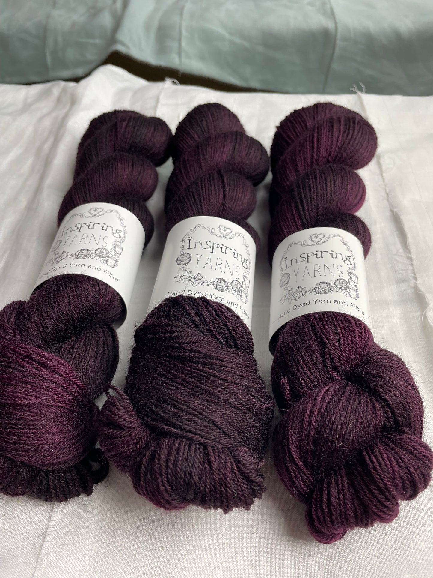 Three skeins of dark burgundy coloured Highland 4Ply labelled Inspiring Yarns on a white linen background.