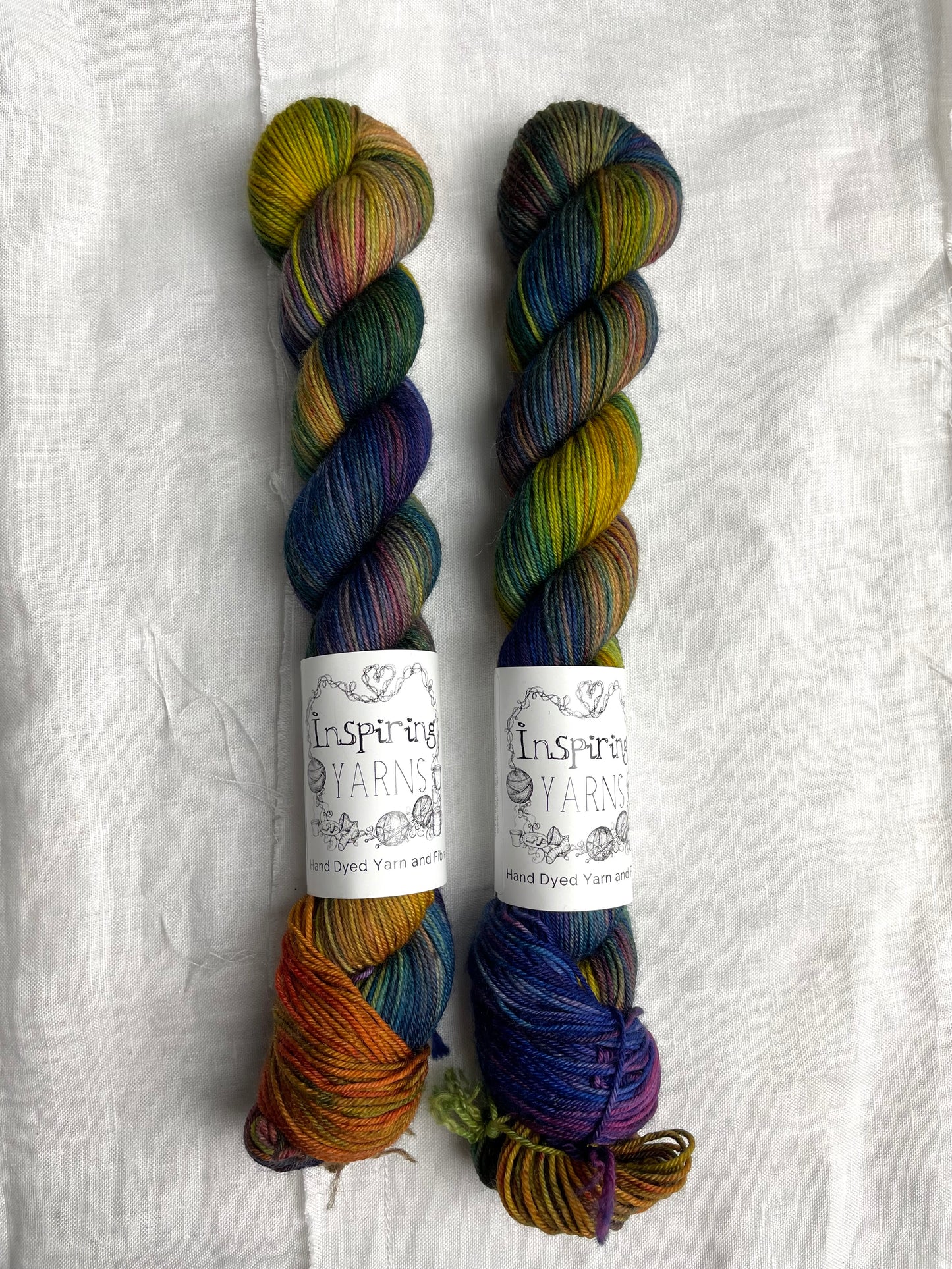 Two skeins of multi coloured Merino Smooth Sock yarn labelled Inspiring Yarns on a white linen background.