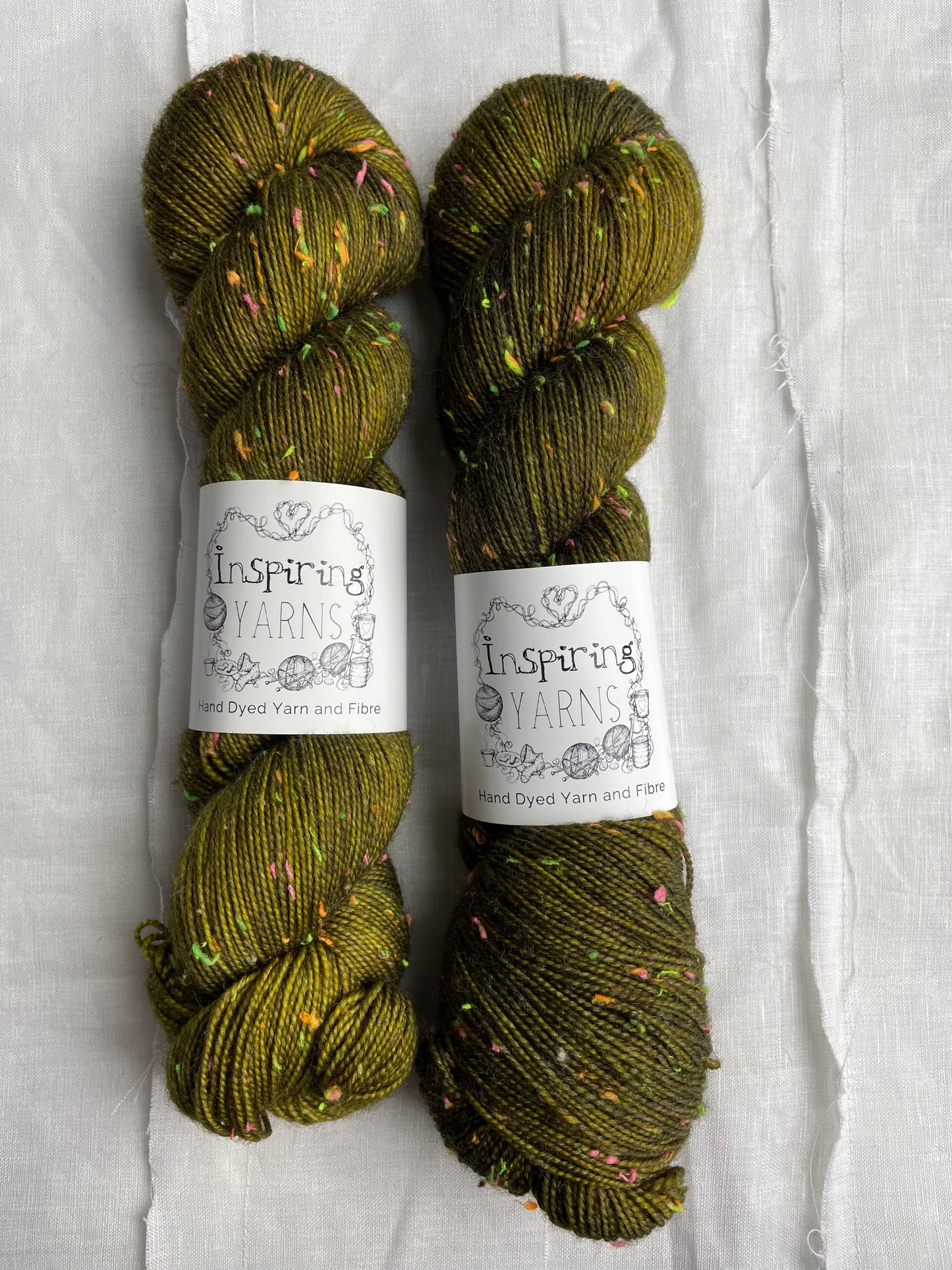Two skeins of tonal dark olive green colour hand dyed Disco Donegal yarn branded Inspiring Yarns on a white linen background. The yarn has multi coloured neon neps.