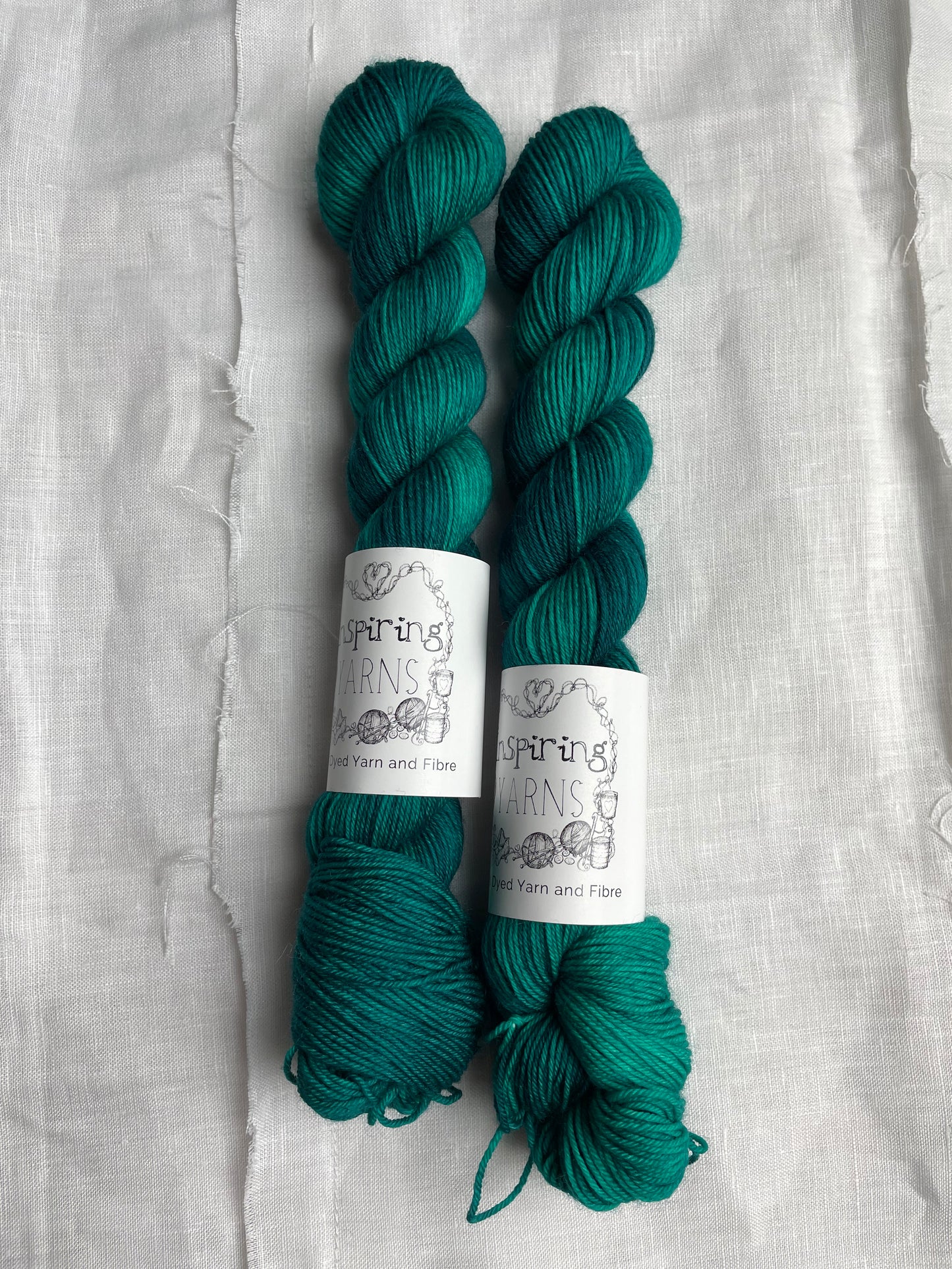 Two skeins of Merino Smooth Sock yarn hand dyed in a tonal jade green colourway and labelled Inspiring Yarns on a white linen background.