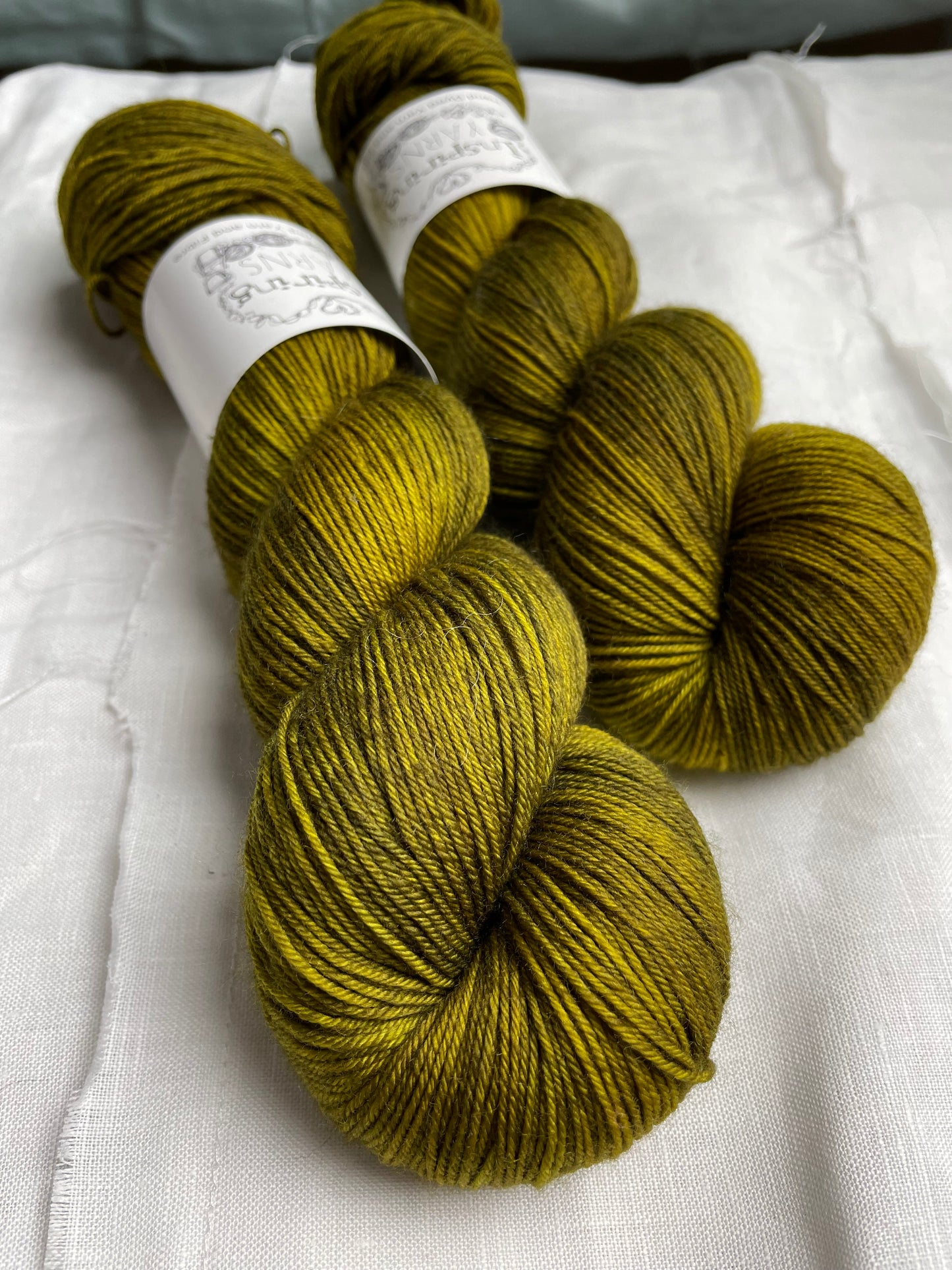 Two skeins of Gremlin, a dark olive green coloured Merino Smooth Sock, labelled Inspiring Yarns on a white linen background.