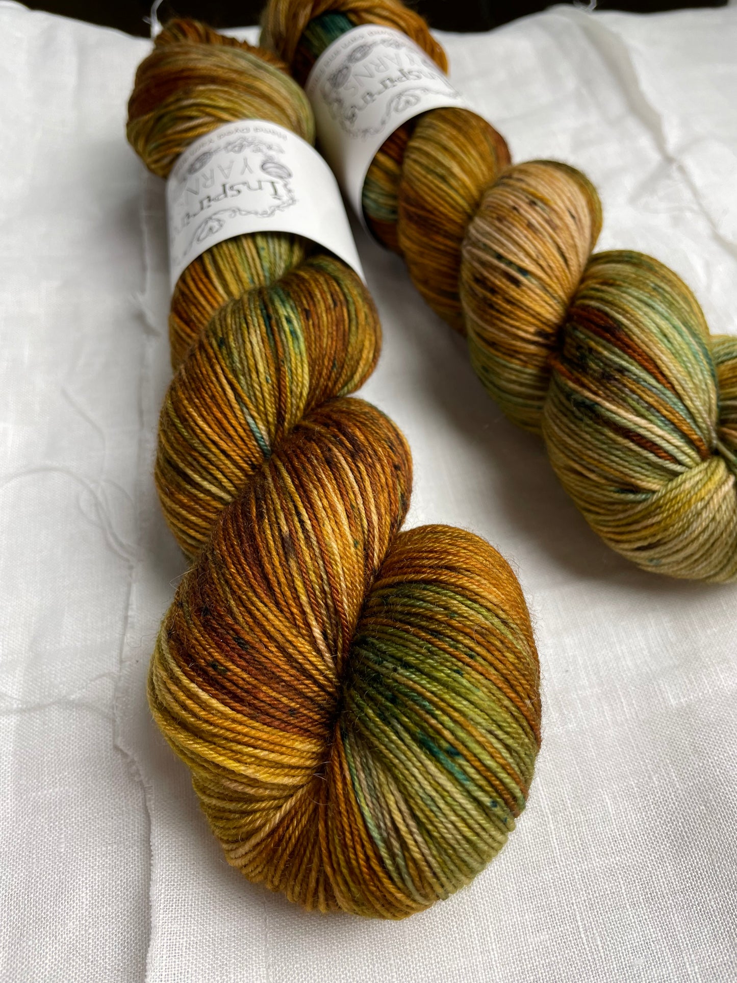 Two skeins of Get Cosy, variegated shades of tan with teal speckles coloured Merino Smooth Sock, labelled Inspiring Yarns on a white linen background.