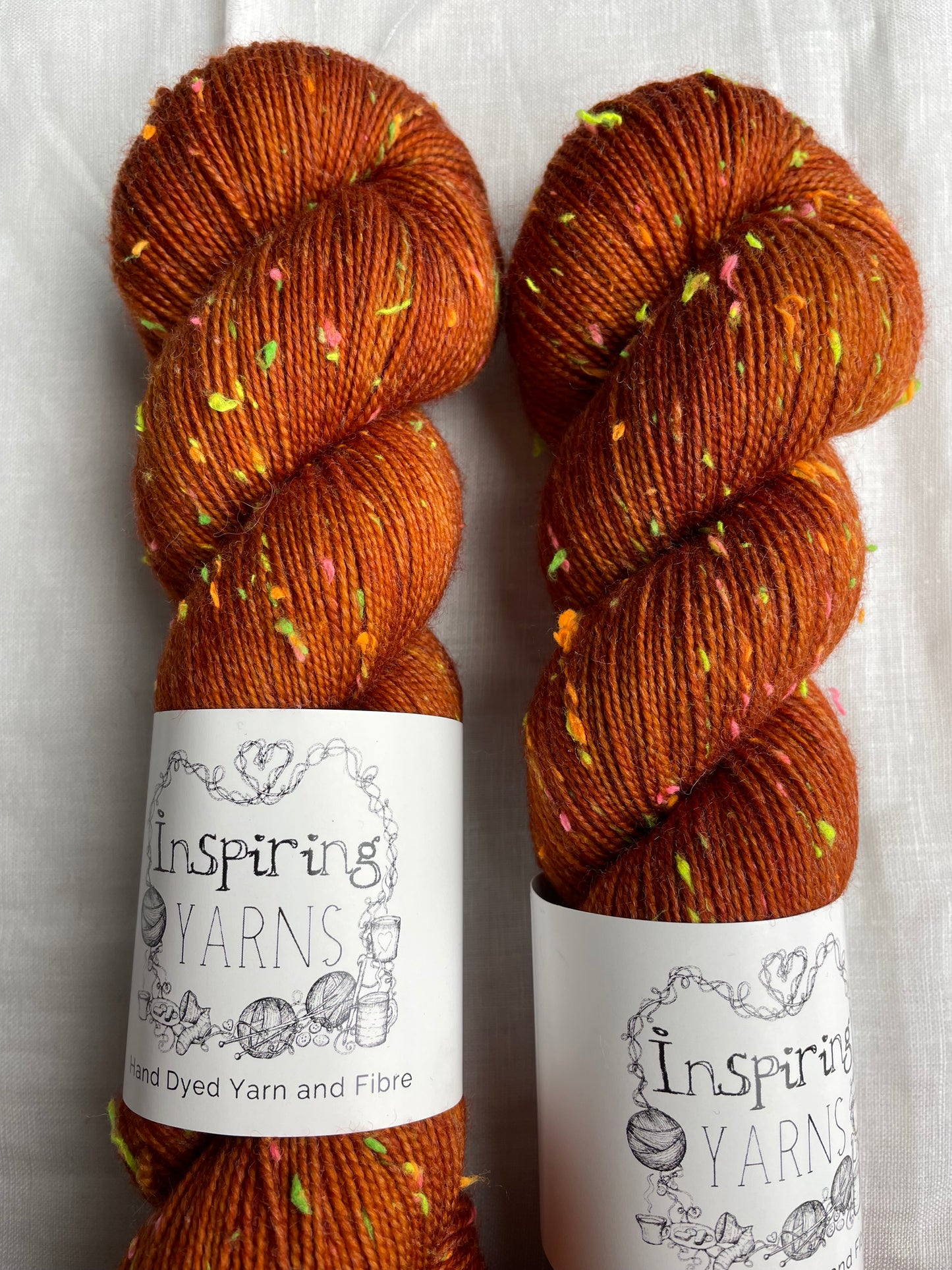 Close up of two skeins of tonal burnt orange coloured hand dyed Disco Donegal yarn branded Inspiring Yarns on a white linen background. The yarn has multi coloured neon neps.